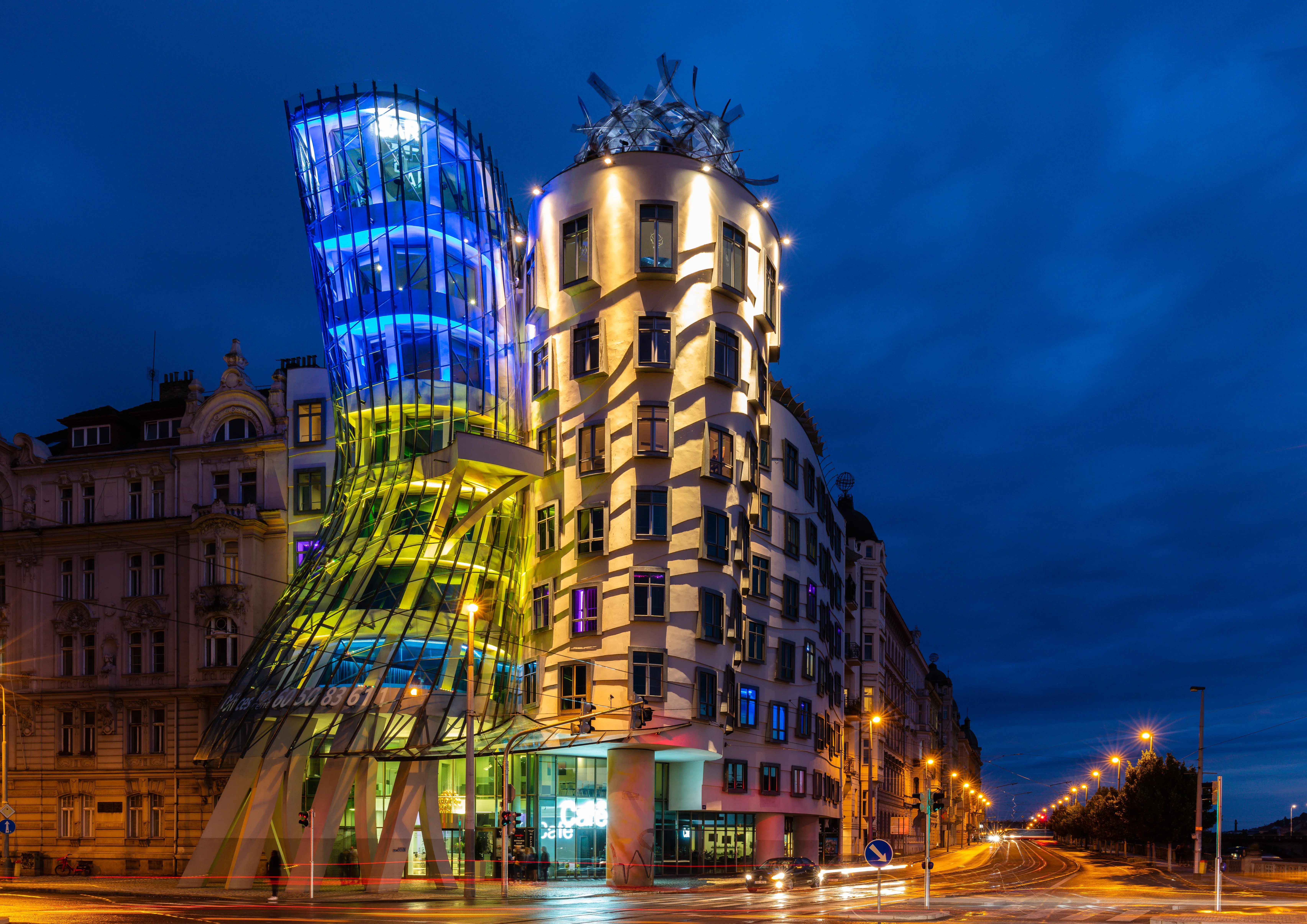La Casa Danzante por la noche, mostrando un edificio moderno con una fachada de vidrio curvada iluminada en azul y verde, junto a un edificio tradicional bajo un cielo azul oscuro.