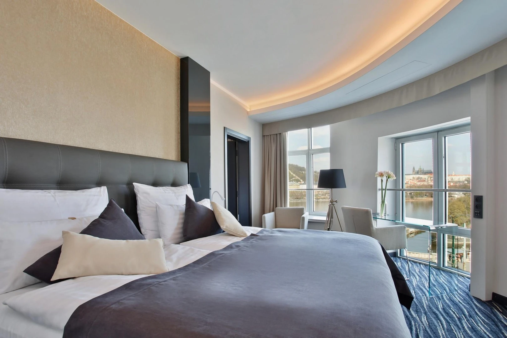 Habitación de hotel moderna con una cama grande, almohadas blancas y oscuras, y vistas a un río a través de una gran ventana.