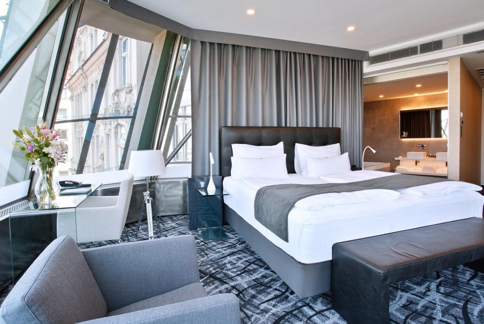 Habitación de hotel moderna con una cama grande, sillón gris y una gran ventana angular con vista a la ciudad.