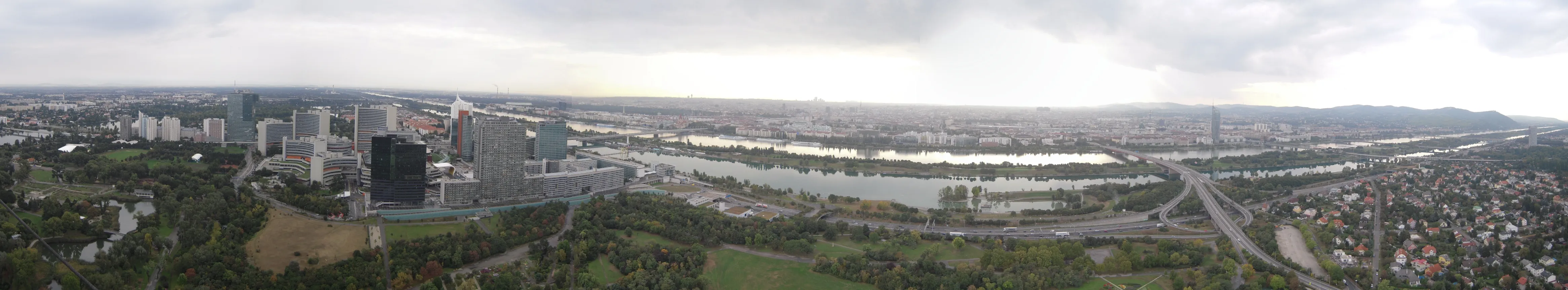 Panoramic cityscape view from a high vantage point showing the Danube River, urban buildings, and green parklands under a cloudy sky