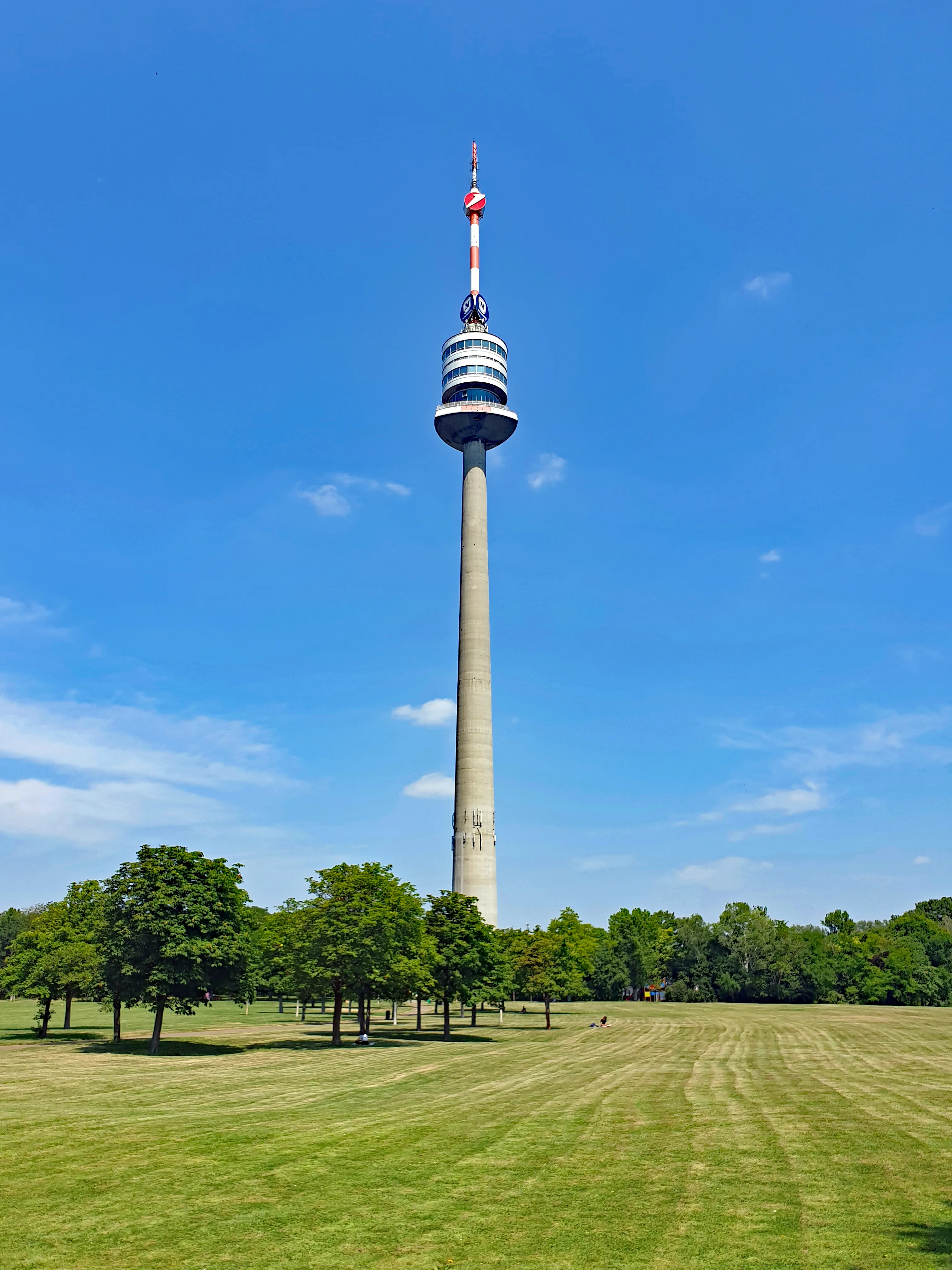 Tall concrete tower with a circular observation deck and red and white antenna against a blue sky, surrounded by green grass and trees