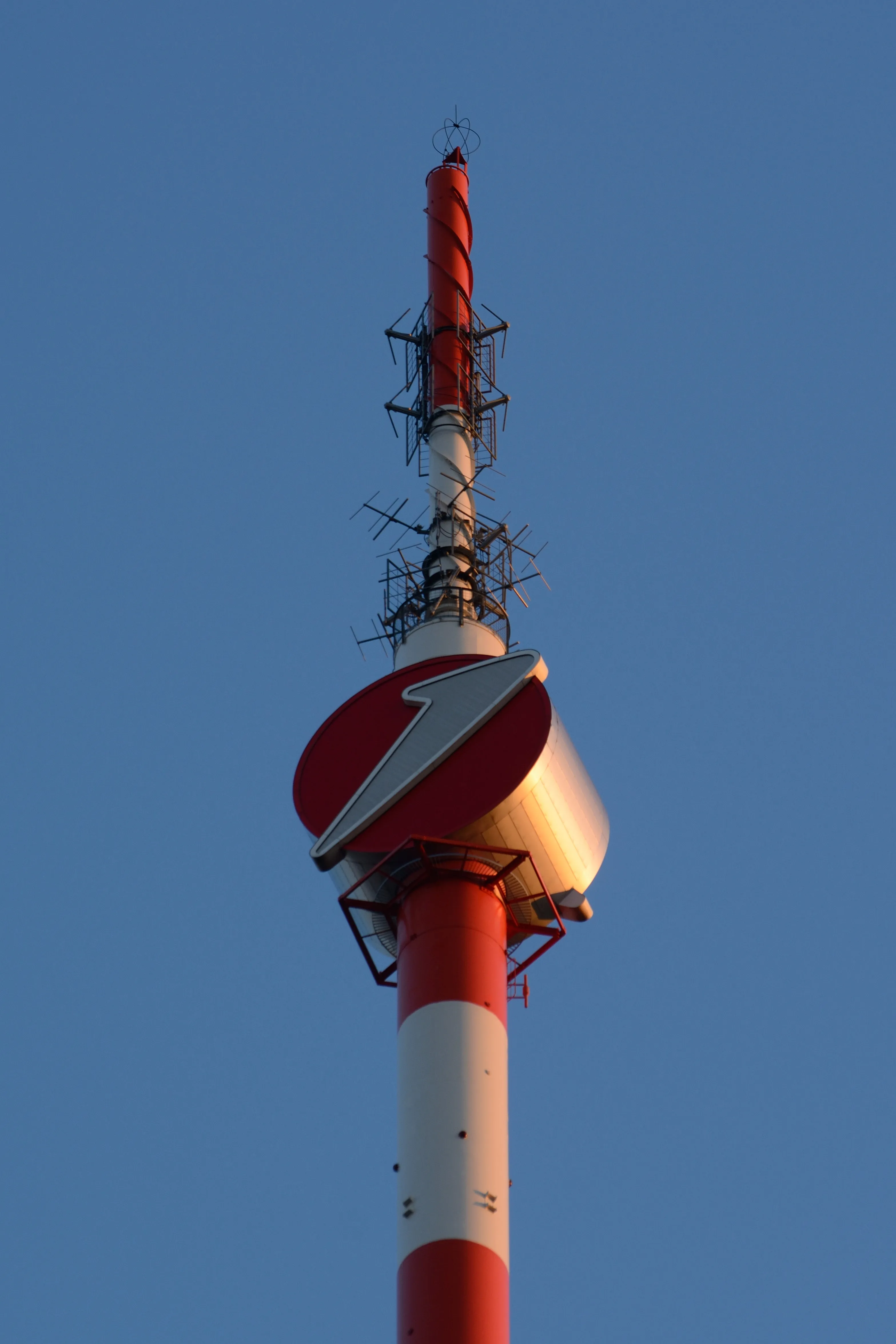 Red and white striped observation tower with a circular sign and antennas against a blue sky
