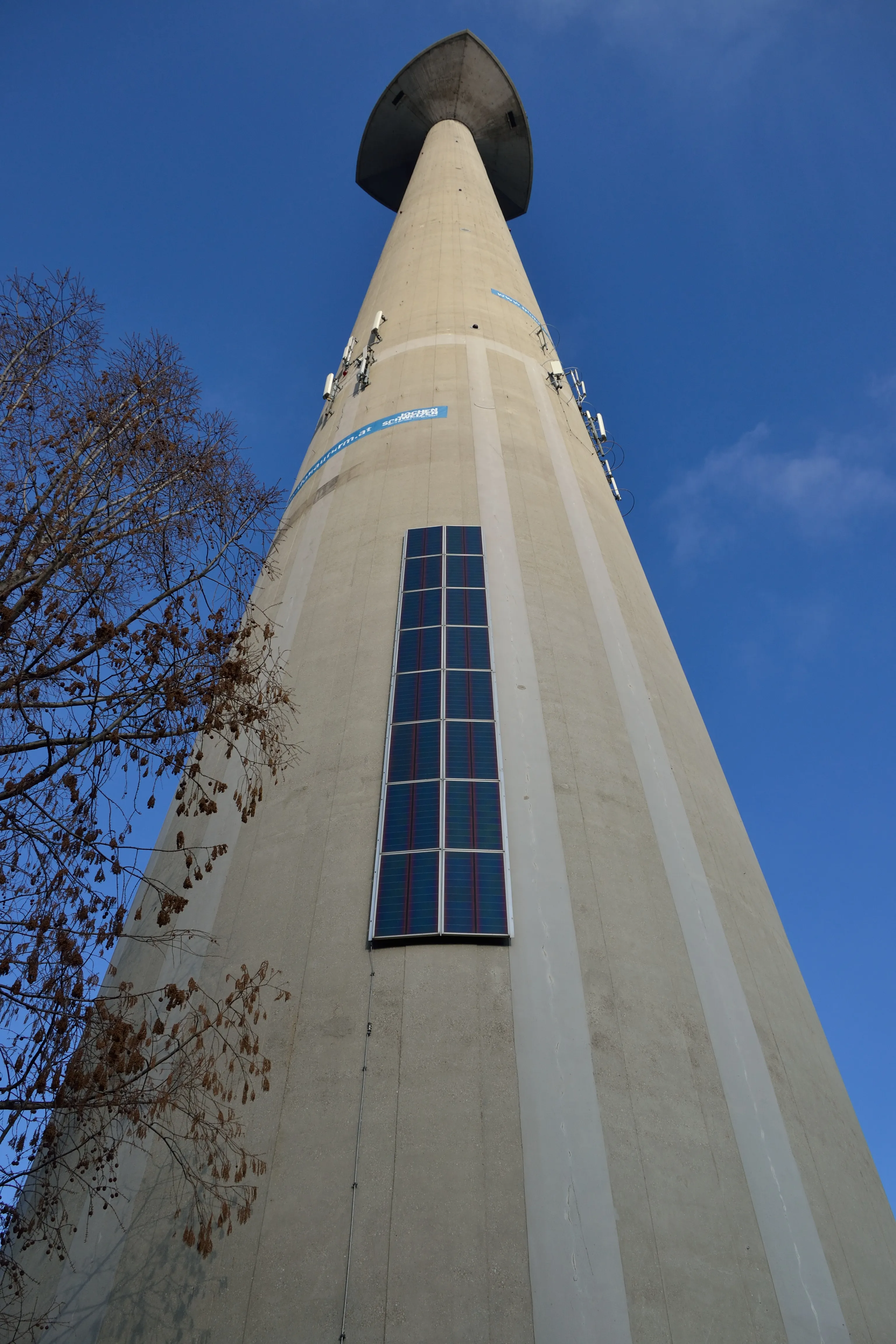 Tall concrete tower with a conical top against a blue sky, featuring a vertical window and communication equipment