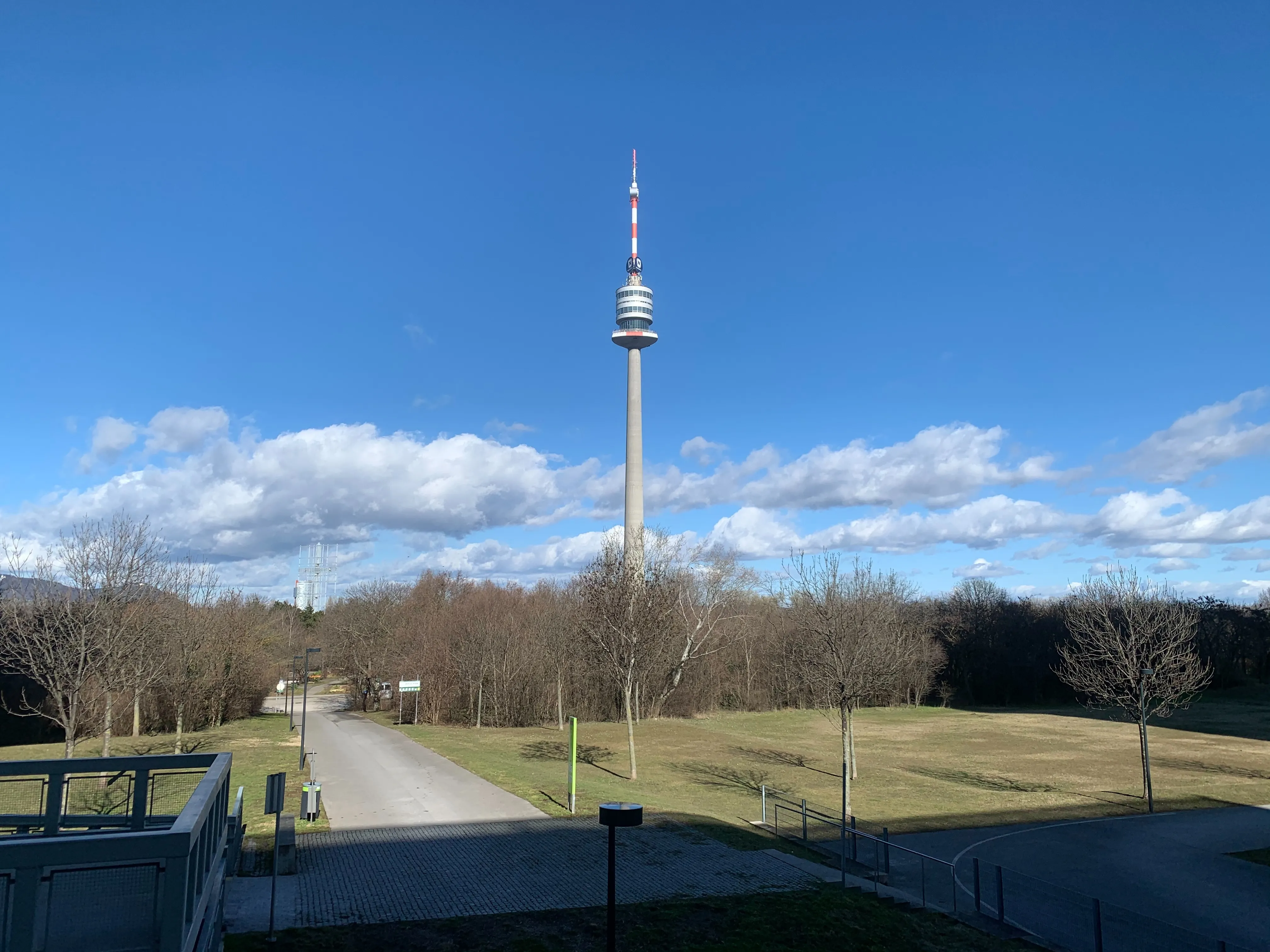 Danube Tower rising above a park with bare trees and a paved path under a blue sky with white clouds