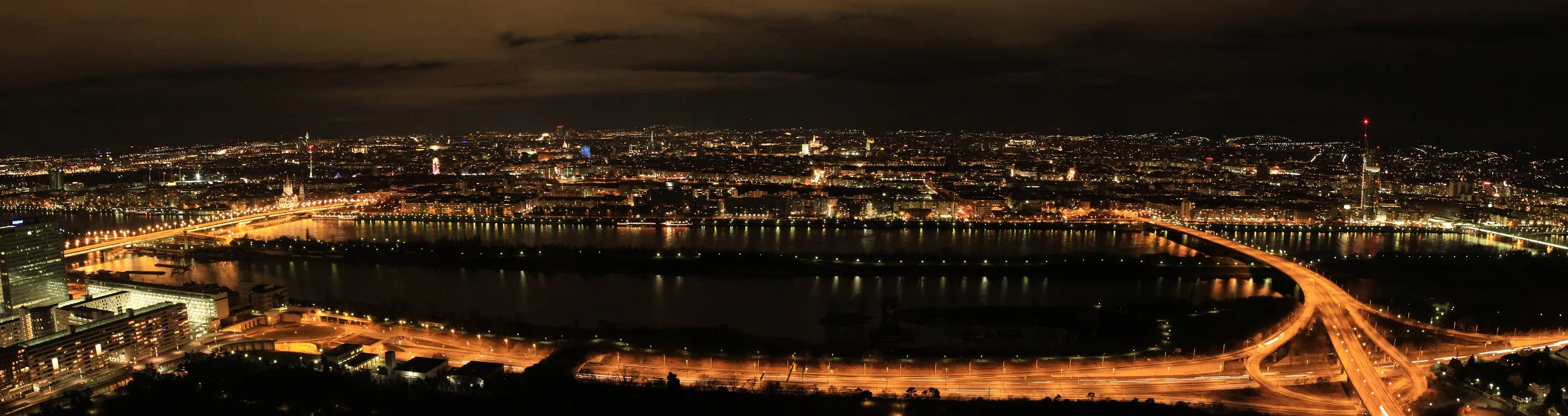 Panoramic night view of Vienna from the Danube Tower, with city lights reflecting on the Danube River and a curved bridge spanning the water.