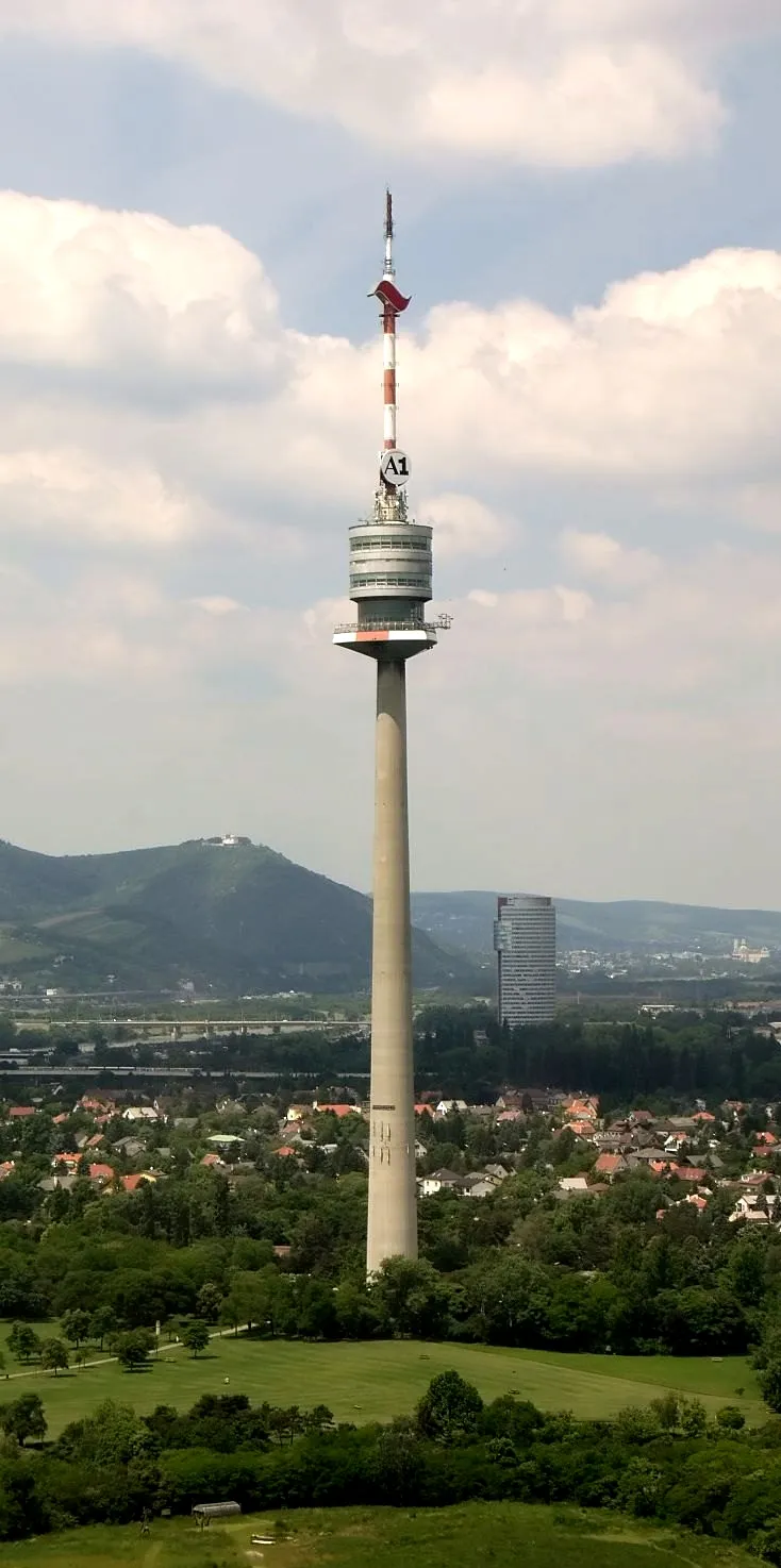 Tall concrete tower with a circular observation deck and red and white antenna spire, surrounded by green parkland and a cityscape with hills in the background
