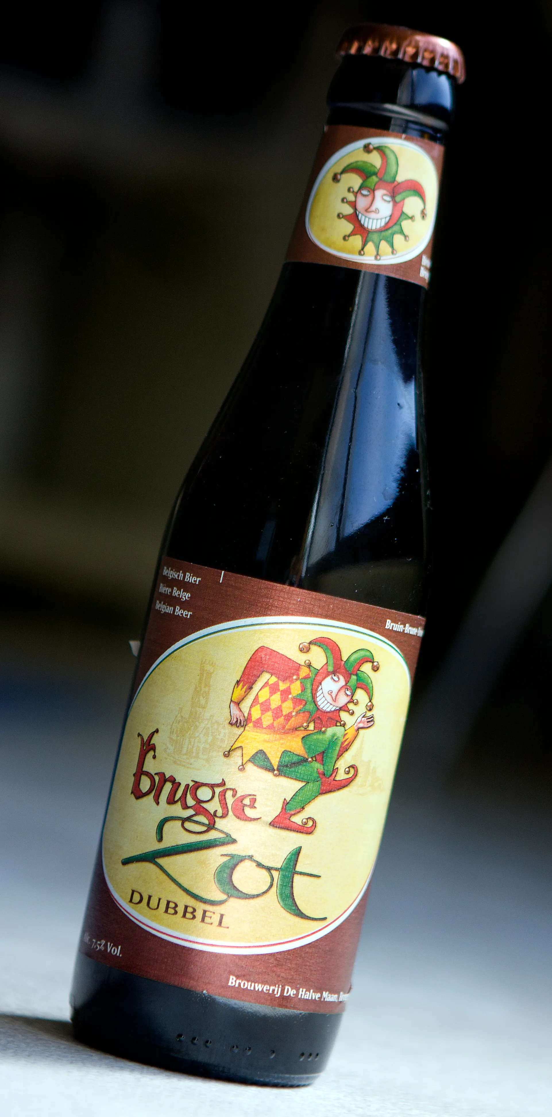 Una botella de cerveza Brugse Zot Dubbel con una etiqueta marrón que presenta una colorida ilustración de un bufón y el nombre Brouwerij De Halve Maan en la parte inferior.