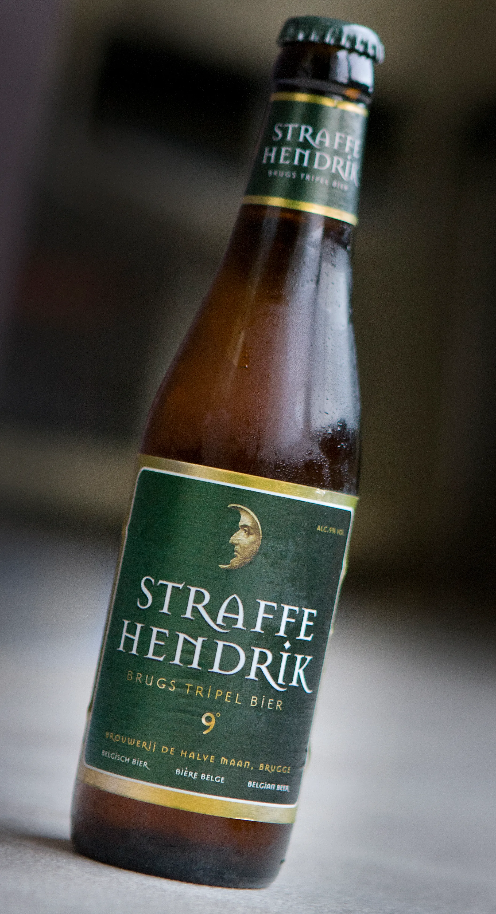 Una botella de cristal marrón de Straffe Hendrik Brugs Tripel Bier con una etiqueta verde oscuro que muestra un logotipo de media luna dorada y un borde dorado, cubierta de gotas de condensación sobre un fondo borroso.