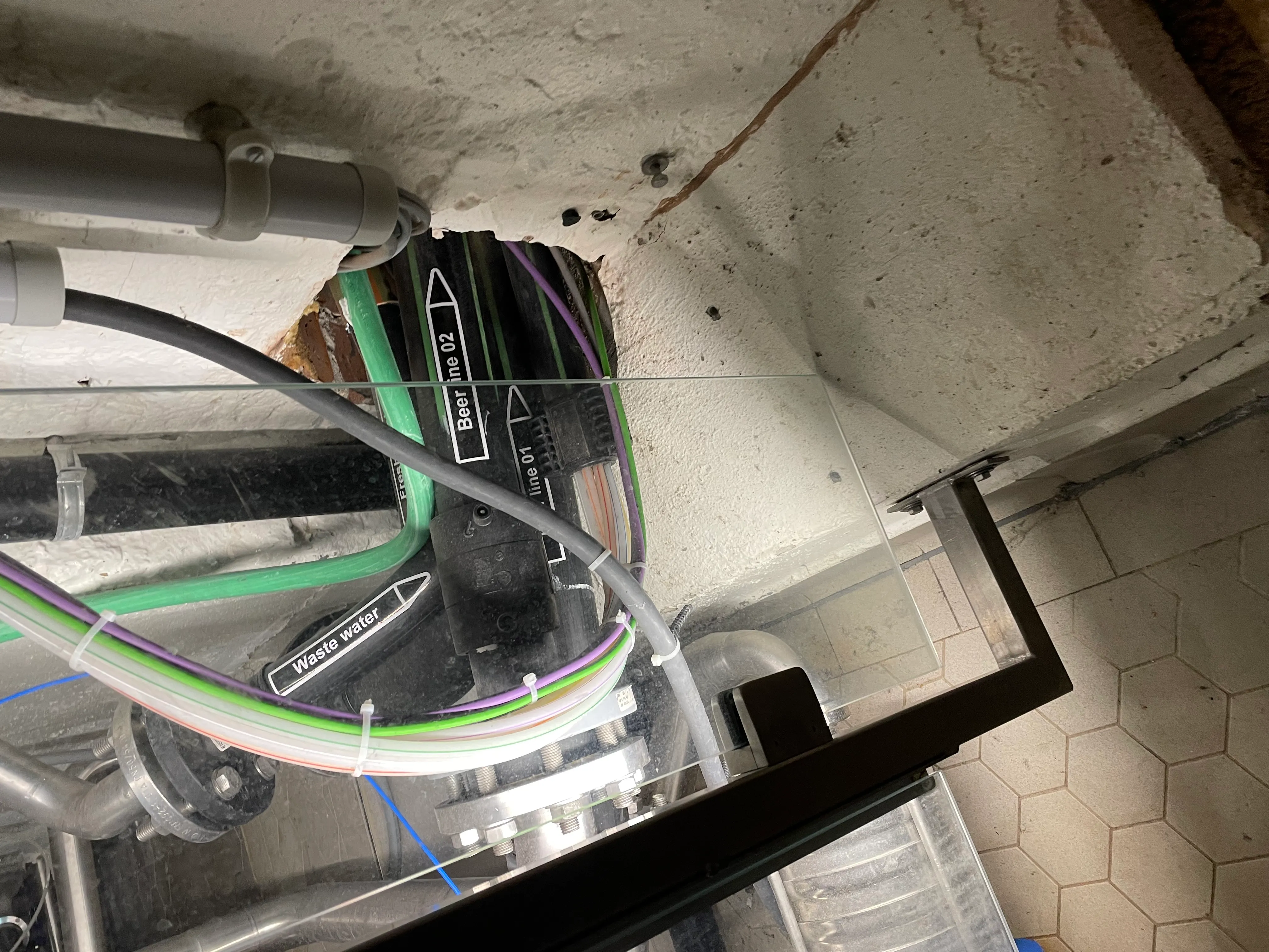 Tubos y cables de colores que atraviesan una abertura de hormigón en el suelo, parte del conducto de cerveza subterráneo de la cervecería De Halve Maan