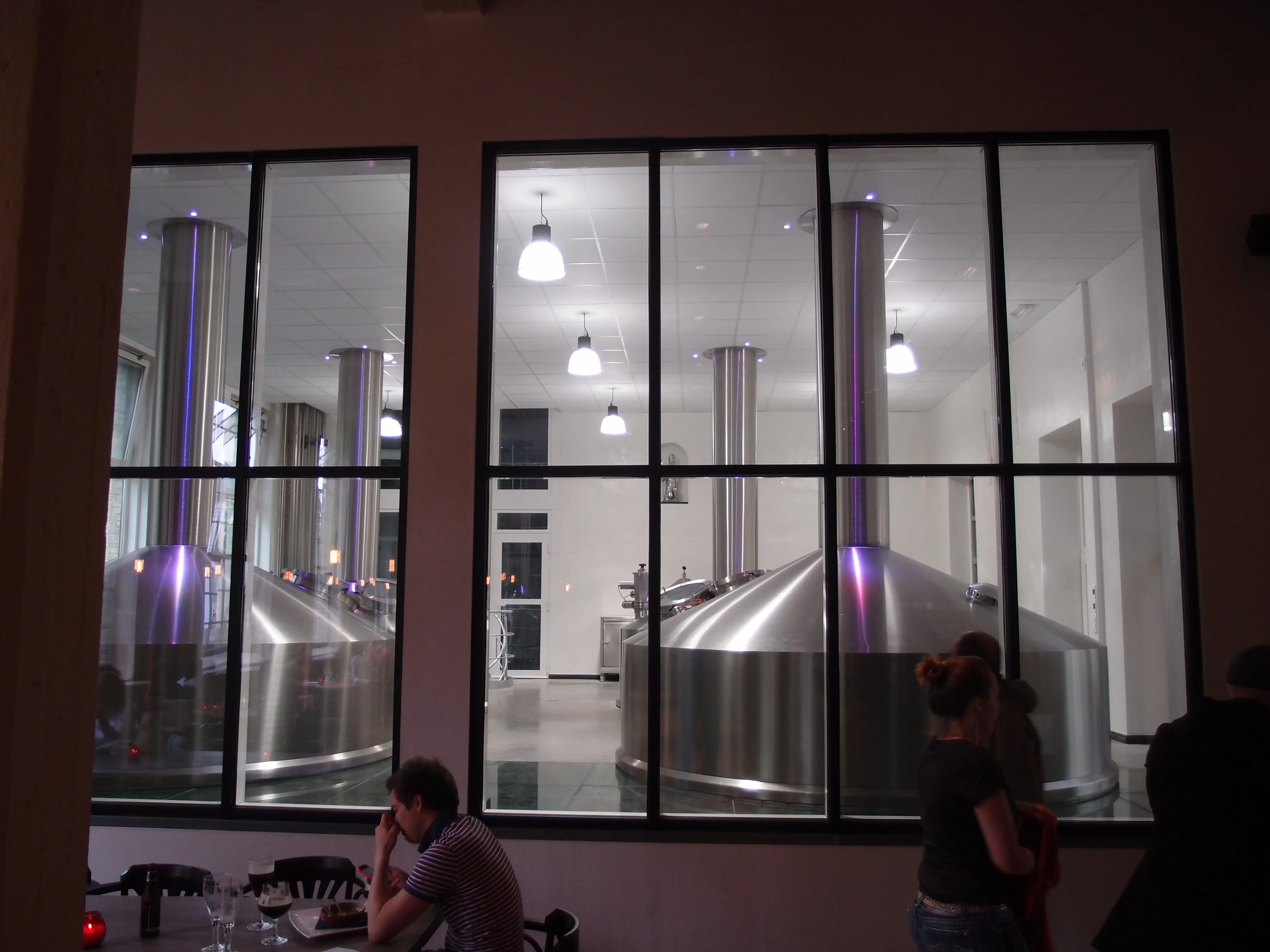 Vista a través de grandes ventanales con marcos negros hacia una sala de elaboración de cerveza con grandes tanques de fermentación de acero inoxidable, con visitantes sentados y de pie en una zona de degustación en primer plano con iluminación tenue.