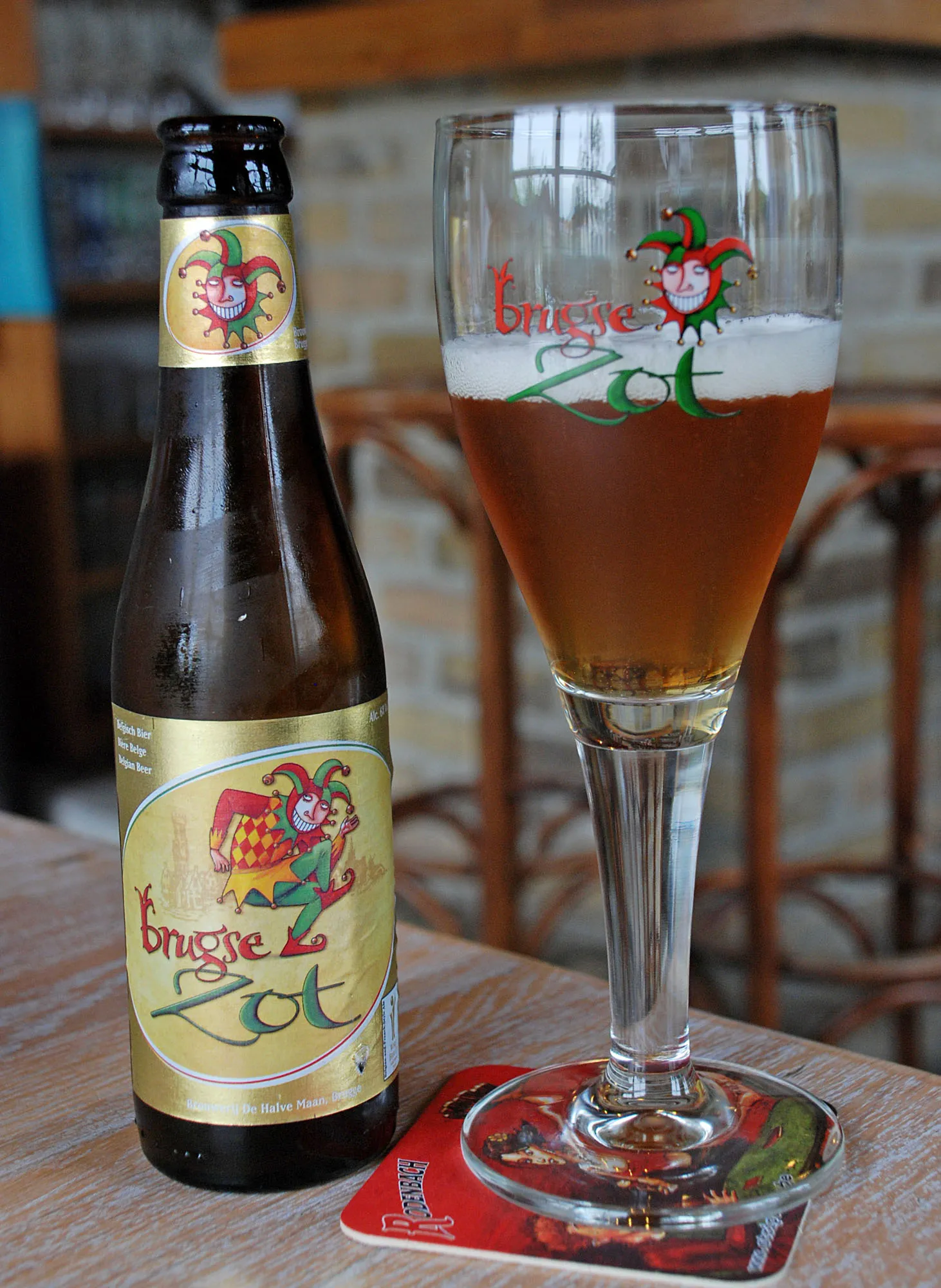 Una botella de vidrio marrón y una copa de tulipa llena de cerveza Brugse Zot con logotipos de bufón sobre una mesa de madera, con un posavasos rojo debajo de la copa.