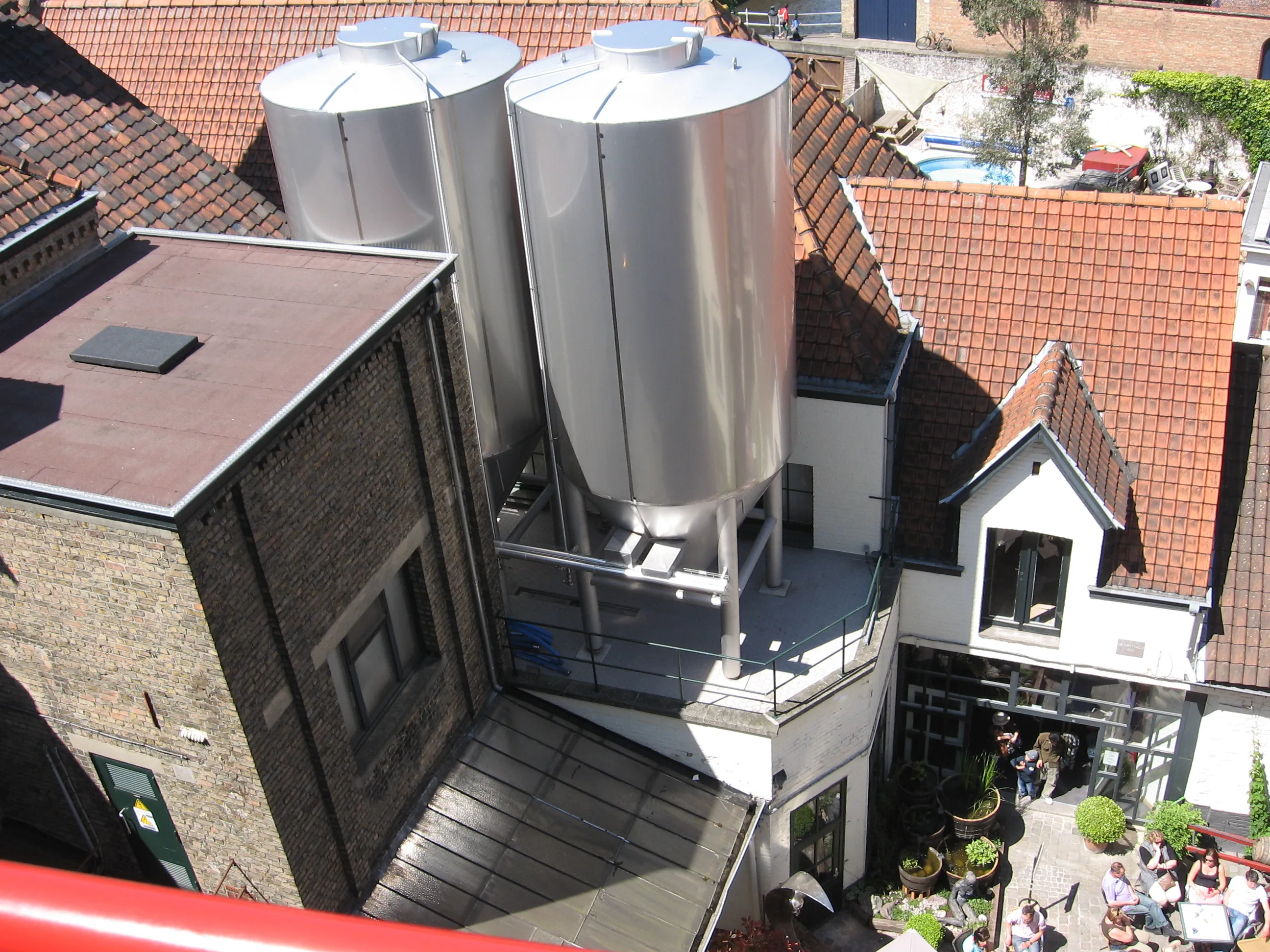 Vista aérea de la Brouwerij De Halve Maan mostrando grandes tanques de fermentación de acero inoxidable en el techo de edificios de ladrillo con tejados rojos y un patio con gente sentada abajo.