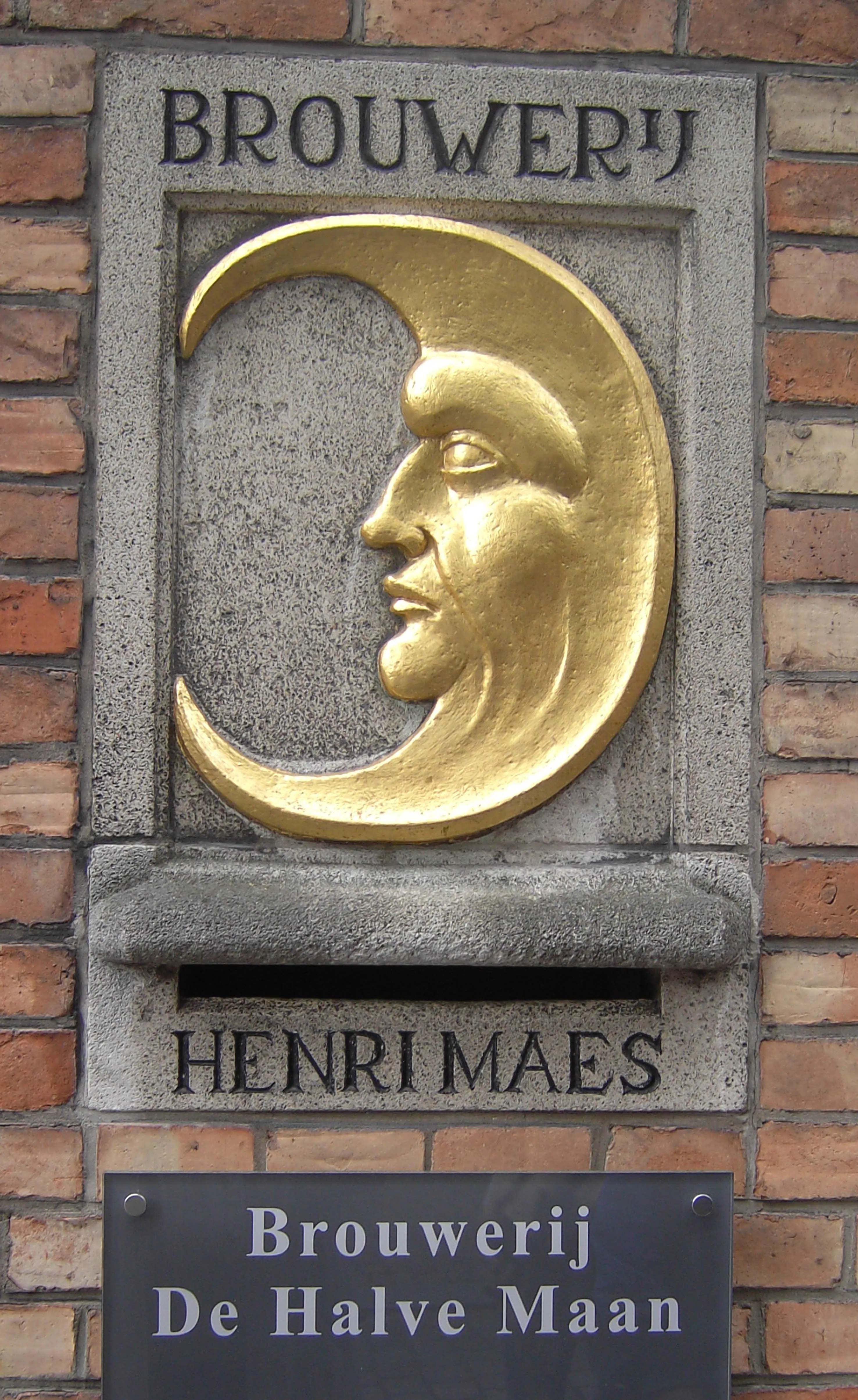 Placa de piedra sobre una pared de ladrillo con una escultura de una media luna dorada con rostro, con la inscripción 'BROUWERIJ' y 'HENRI MAES', sobre un cartel que reza 'Brouwerij De Halve Maan'.