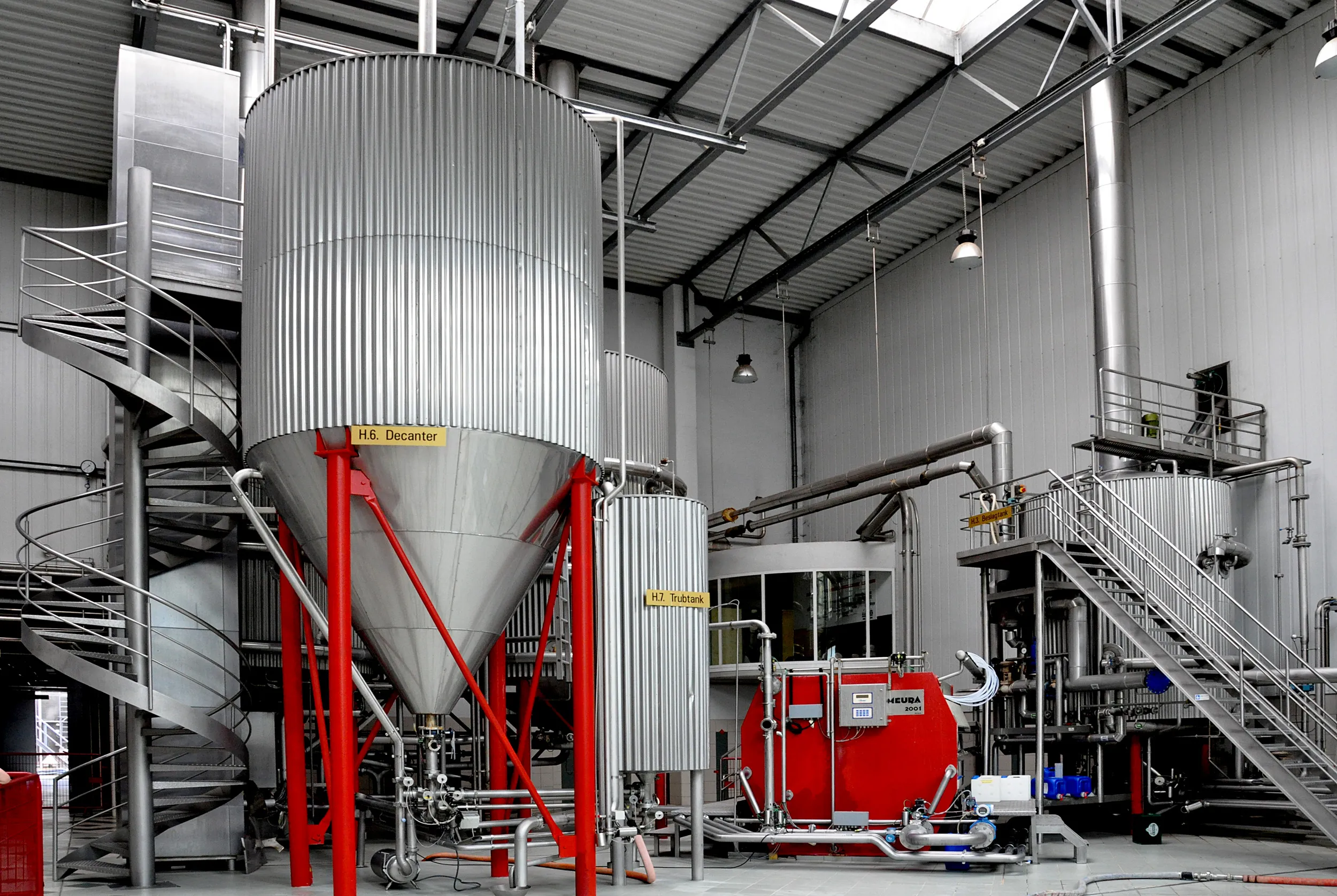 Grandes tanques de fermentación de acero inoxidable con estructuras de soporte rojas y escaleras de metal dentro de la sala de producción de la cervecería De Koninck
