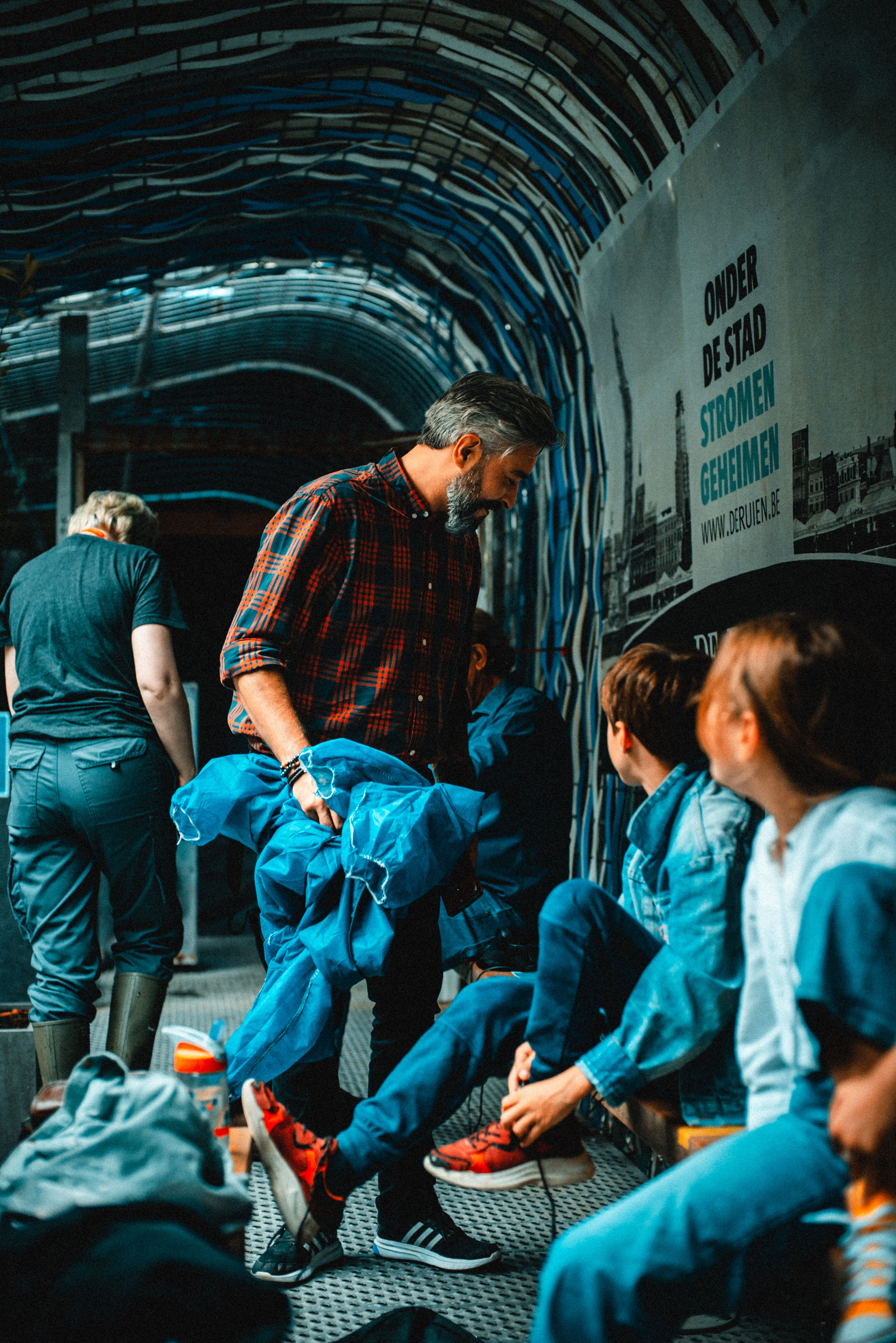 Grupo de adultos y niños con trajes impermeables azules dentro de un túnel subterráneo curvo con tuberías en el techo y un cartel de De Ruien en la pared.