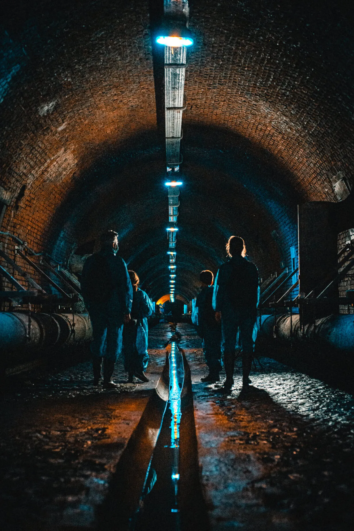 Grupo de personas caminando por un túnel subterráneo oscuro con arcos de ladrillo, un canal de agua central y luces azules en el techo.
