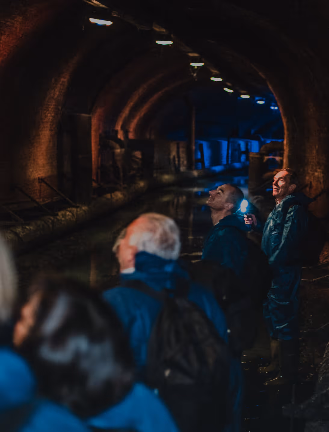 Grupo de visitantes con chaquetas azules de pie en un túnel subterráneo oscuro con arcos de ladrillo, agua y luces en el techo, mirando hacia arriba