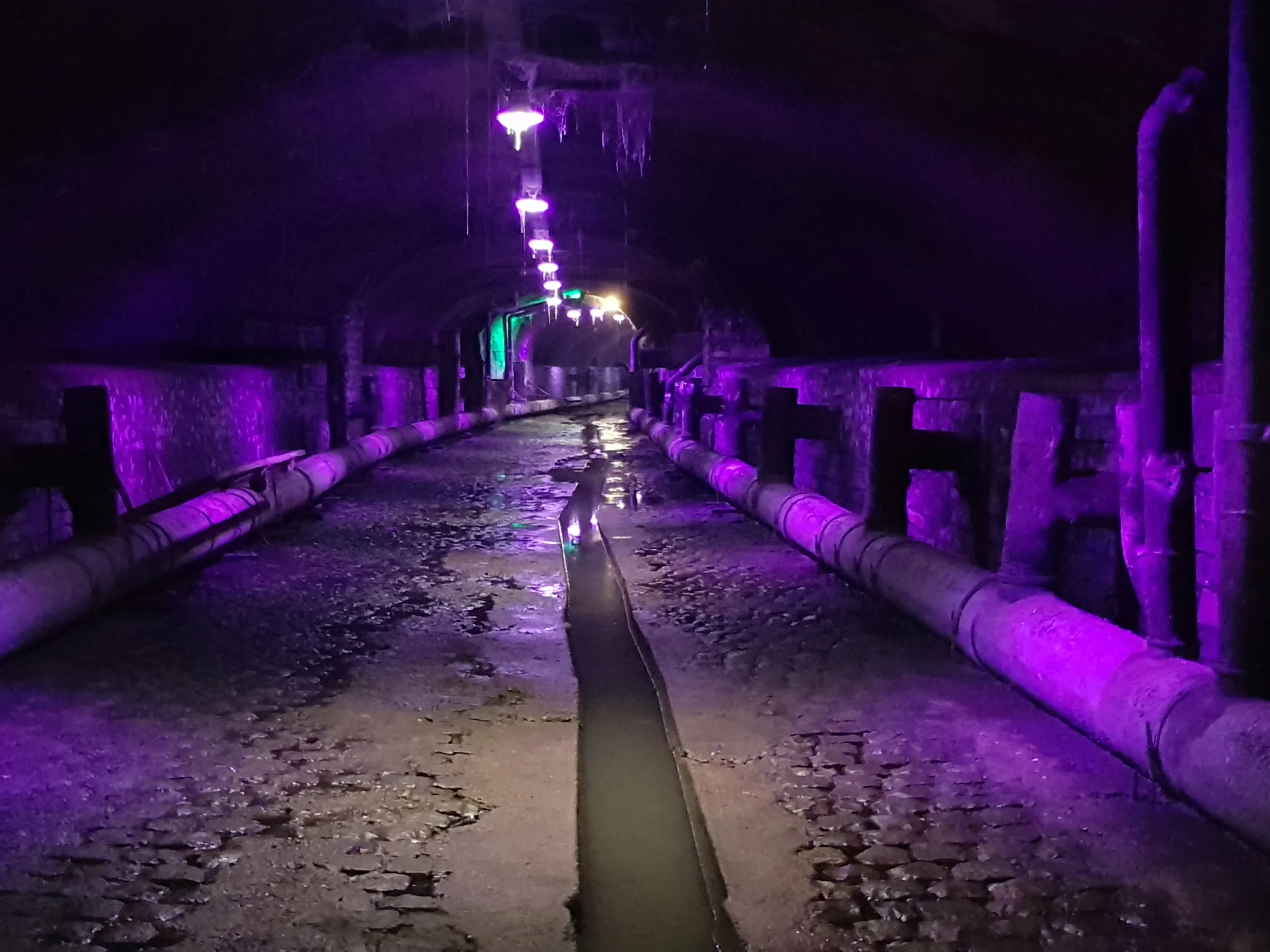 Túnel subterráneo de canal iluminado con luces moradas, que presenta tuberías a lo largo de paredes de ladrillo y un canal de agua que atraviesa el centro
