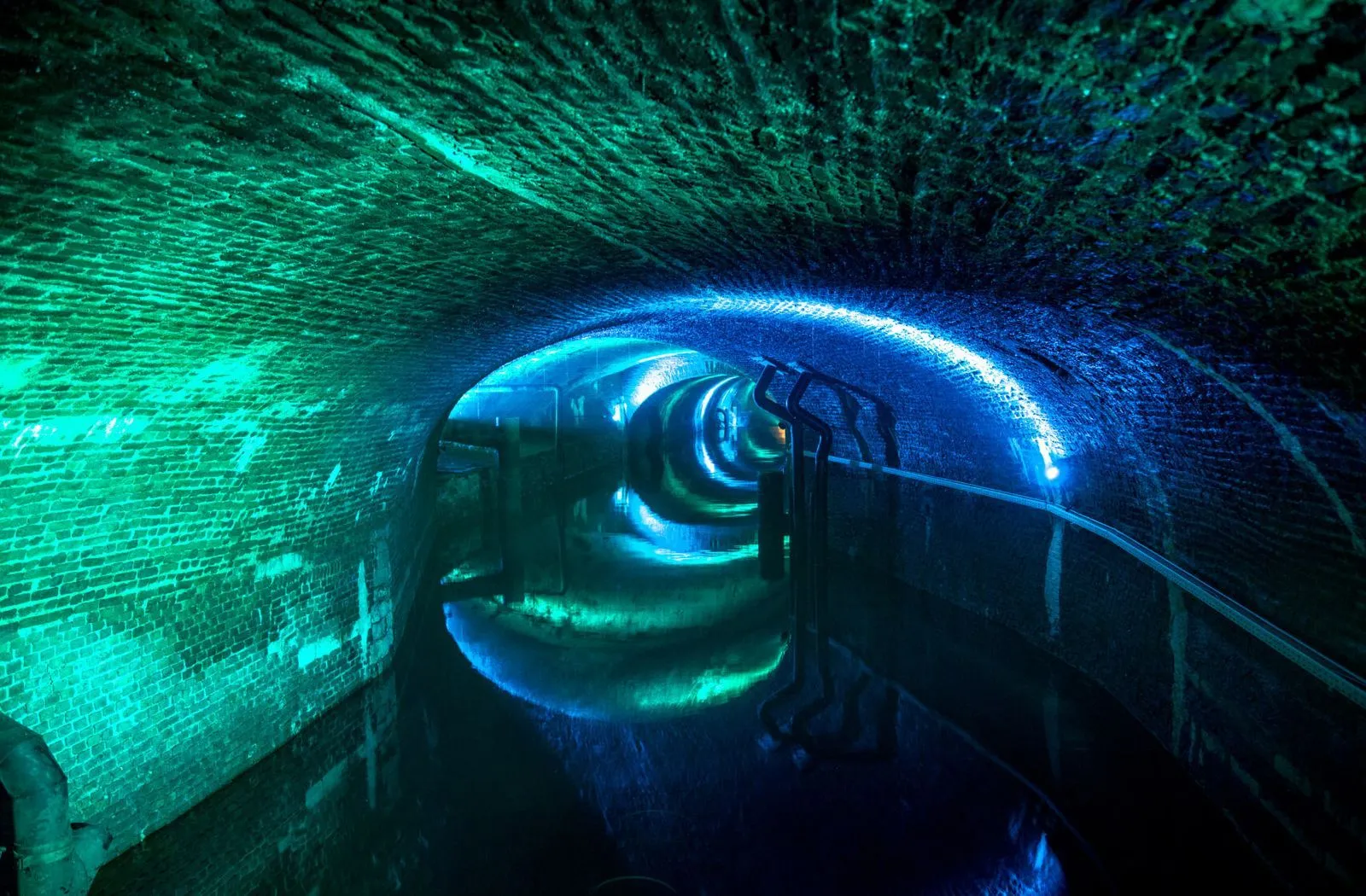 Túnel subterráneo arqueado y revestido de ladrillo, iluminado con luces verdes y azules intensas, que cuenta con una pasarela metálica curva con barandilla a lo largo de un canal de agua oscuro.