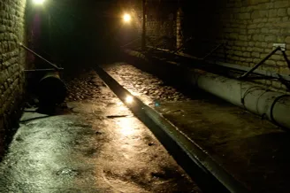 Túnel subterráneo oscuro de ladrillo con un canal de agua en el centro y tuberías montadas a lo largo de la pared derecha, iluminado por tenues luces de techo.