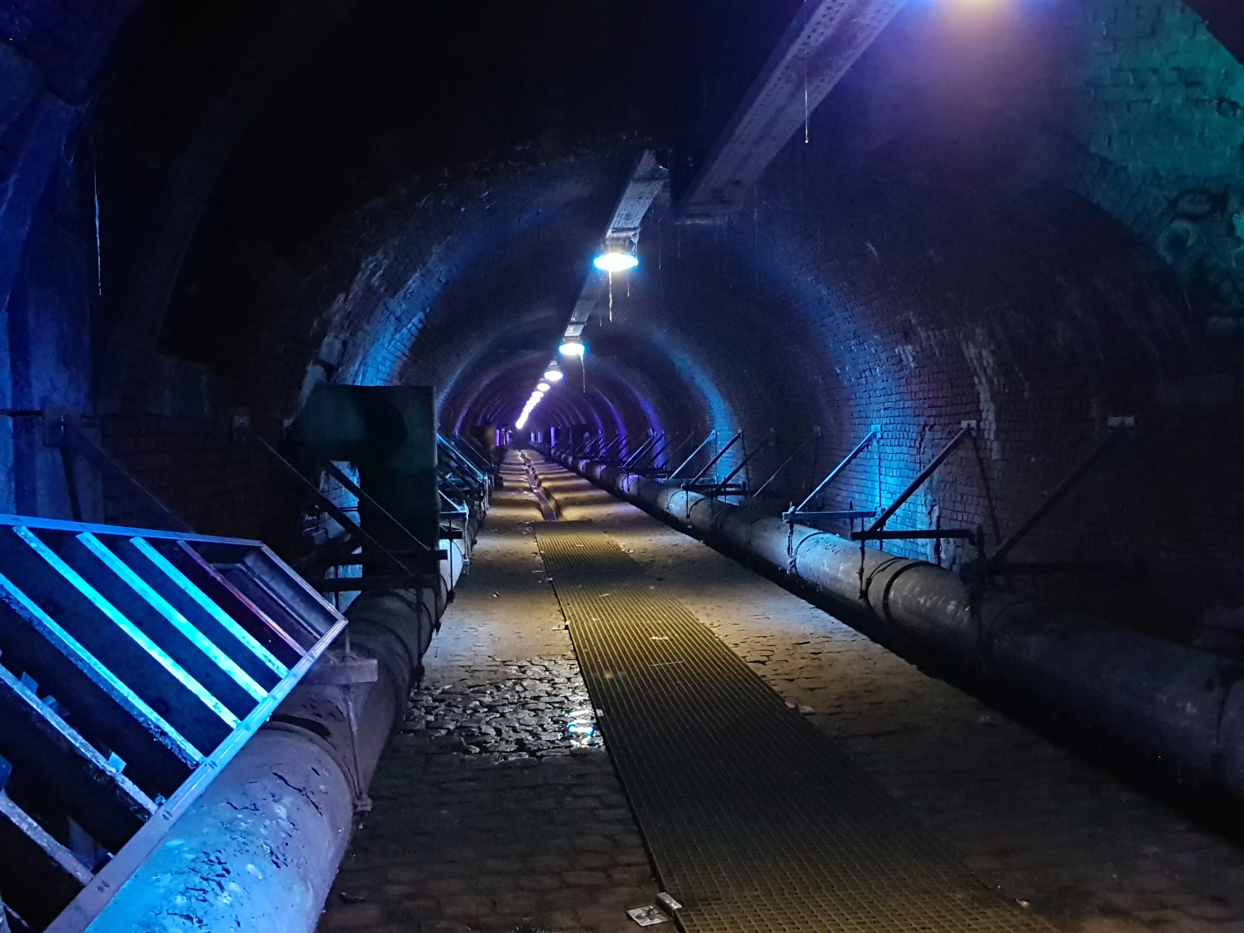Largo túnel de ladrillo abovedado con iluminación azul, grandes tuberías a lo largo de ambas paredes y una pasarela central en el sistema de canales subterráneo De Ruien