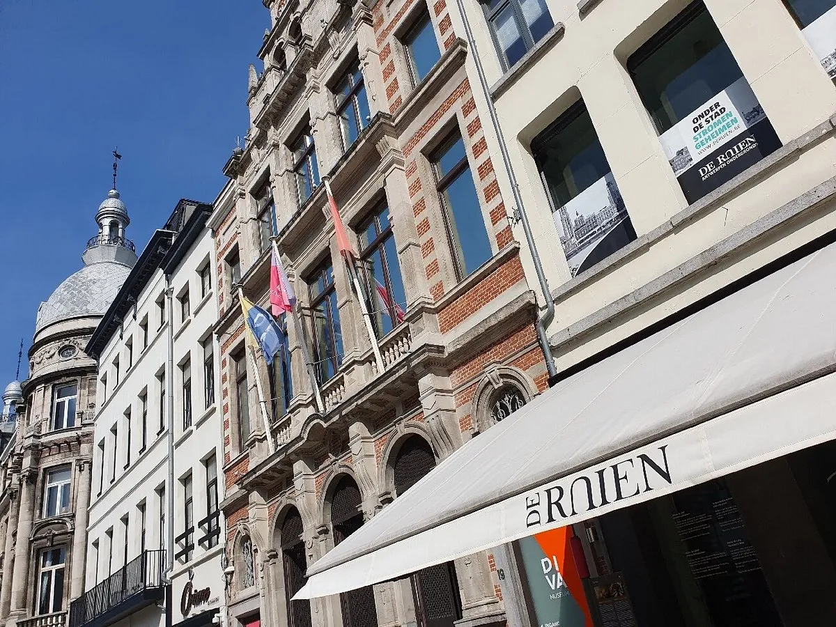 Vista de la calle con edificios históricos de Amberes, un toldo blanco con el letrero 'DE ROIEN' y banderas en una fachada de ladrillo