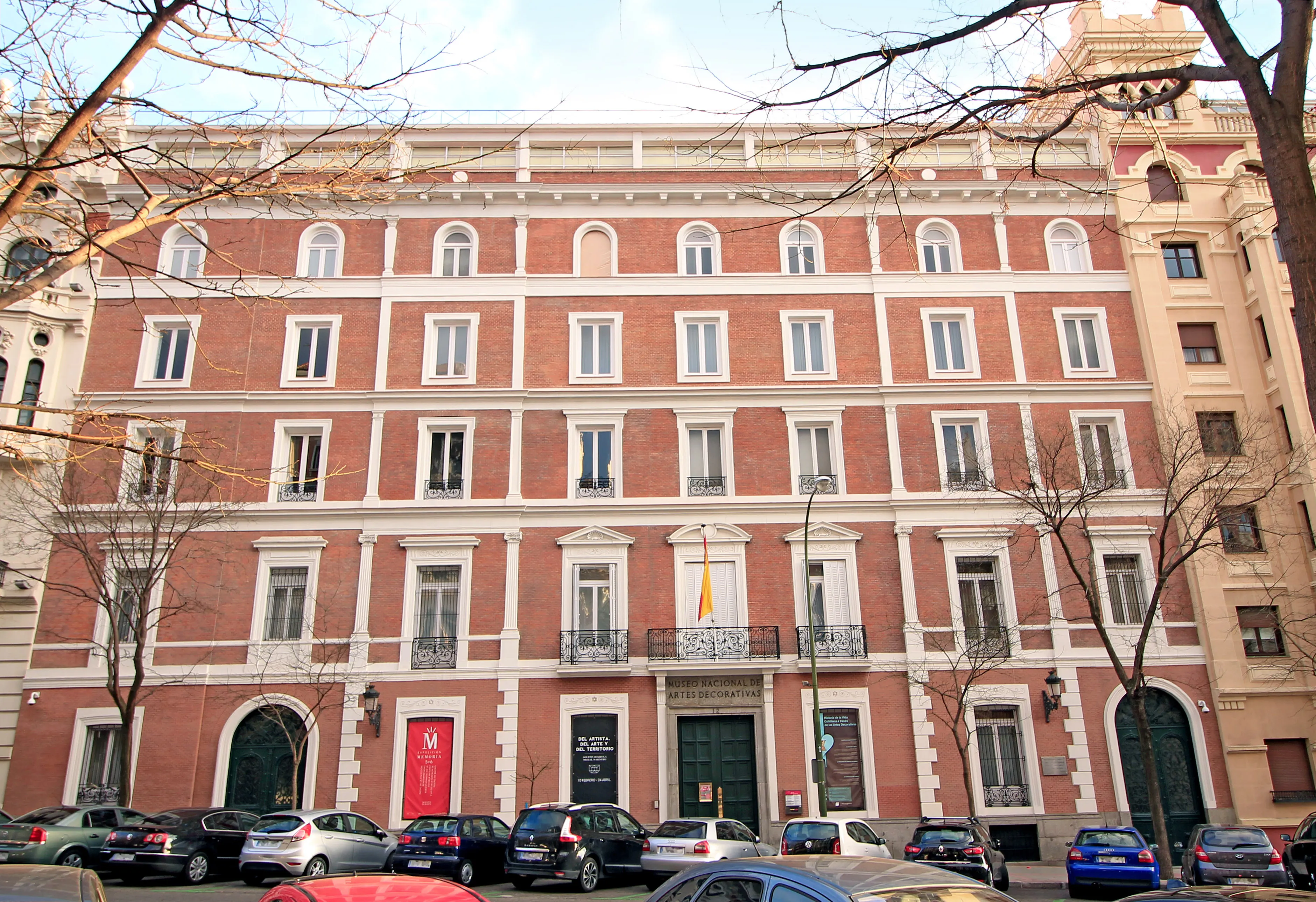 Un gran edificio de ladrillo rojo con detalles blancos y múltiples ventanas, que presenta una entrada central con balcón y una bandera española, rodeado de coches aparcados y árboles desnudos.