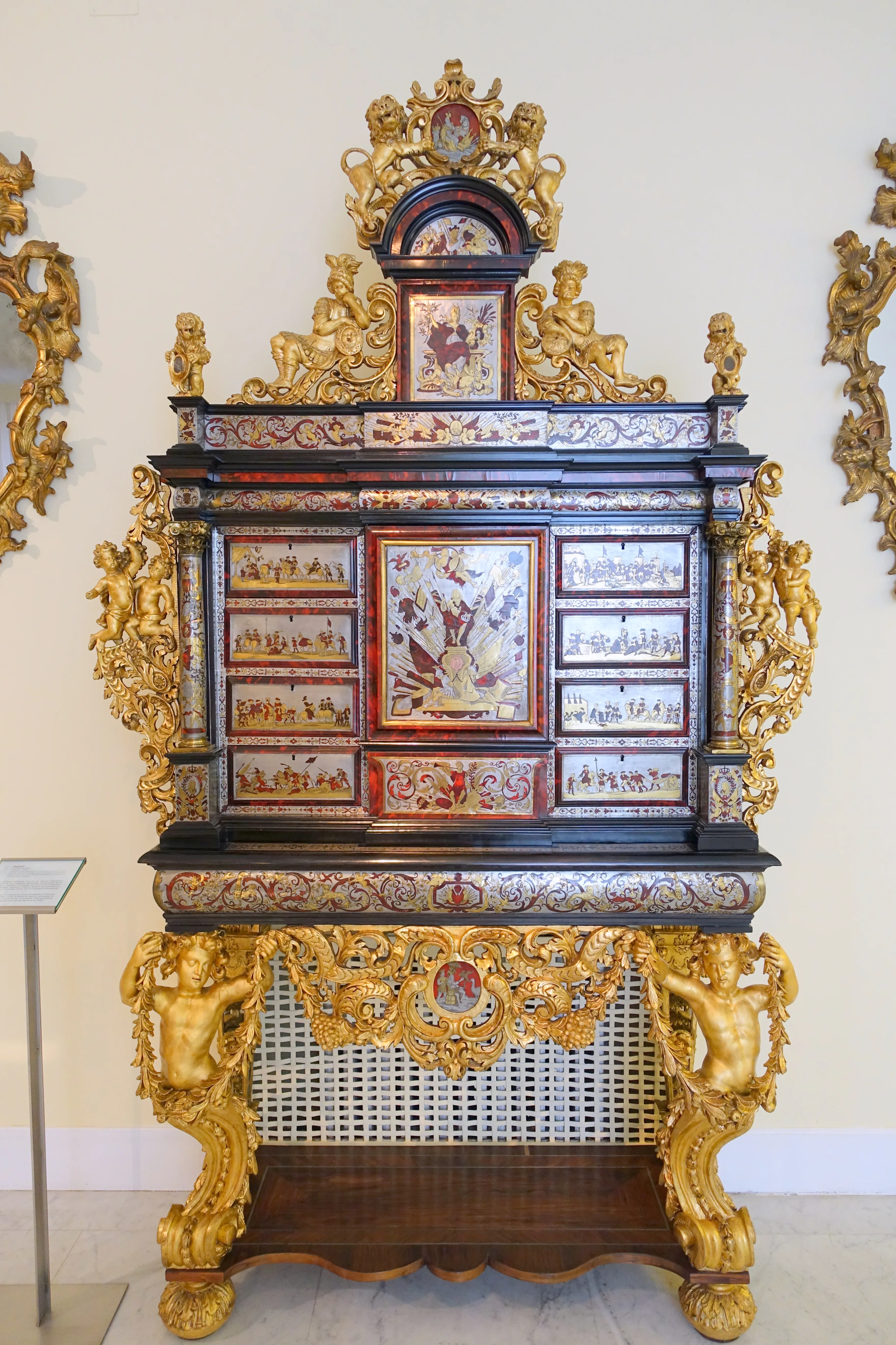 Ornate gilded cabinet with intricate carvings and painted panels displayed in a museum