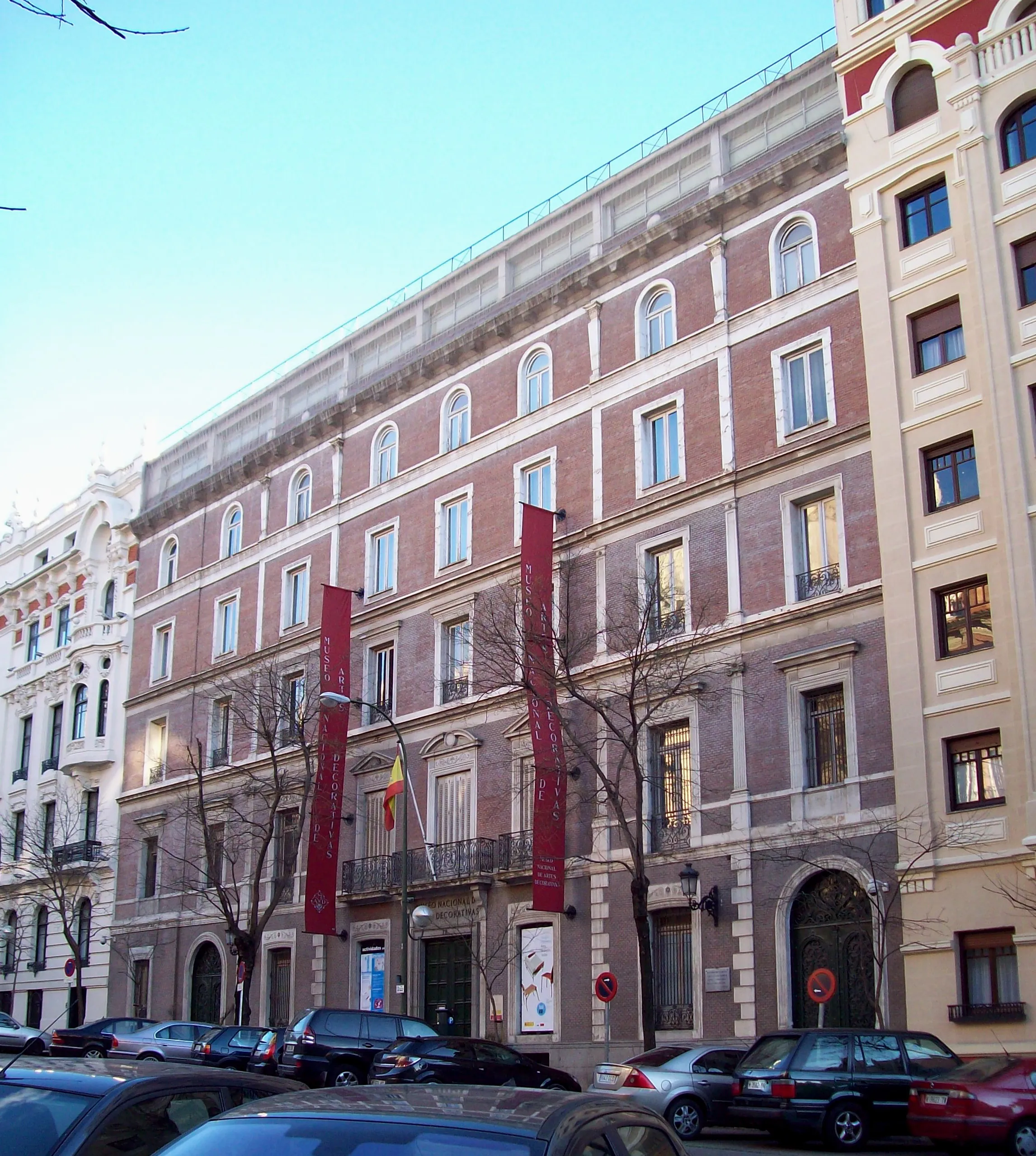 Un edificio de varias plantas con fachada clásica, ventanas de arco y pancartas rojas que cuelgan de los pisos superiores, situado en una calle de la ciudad con coches aparcados.