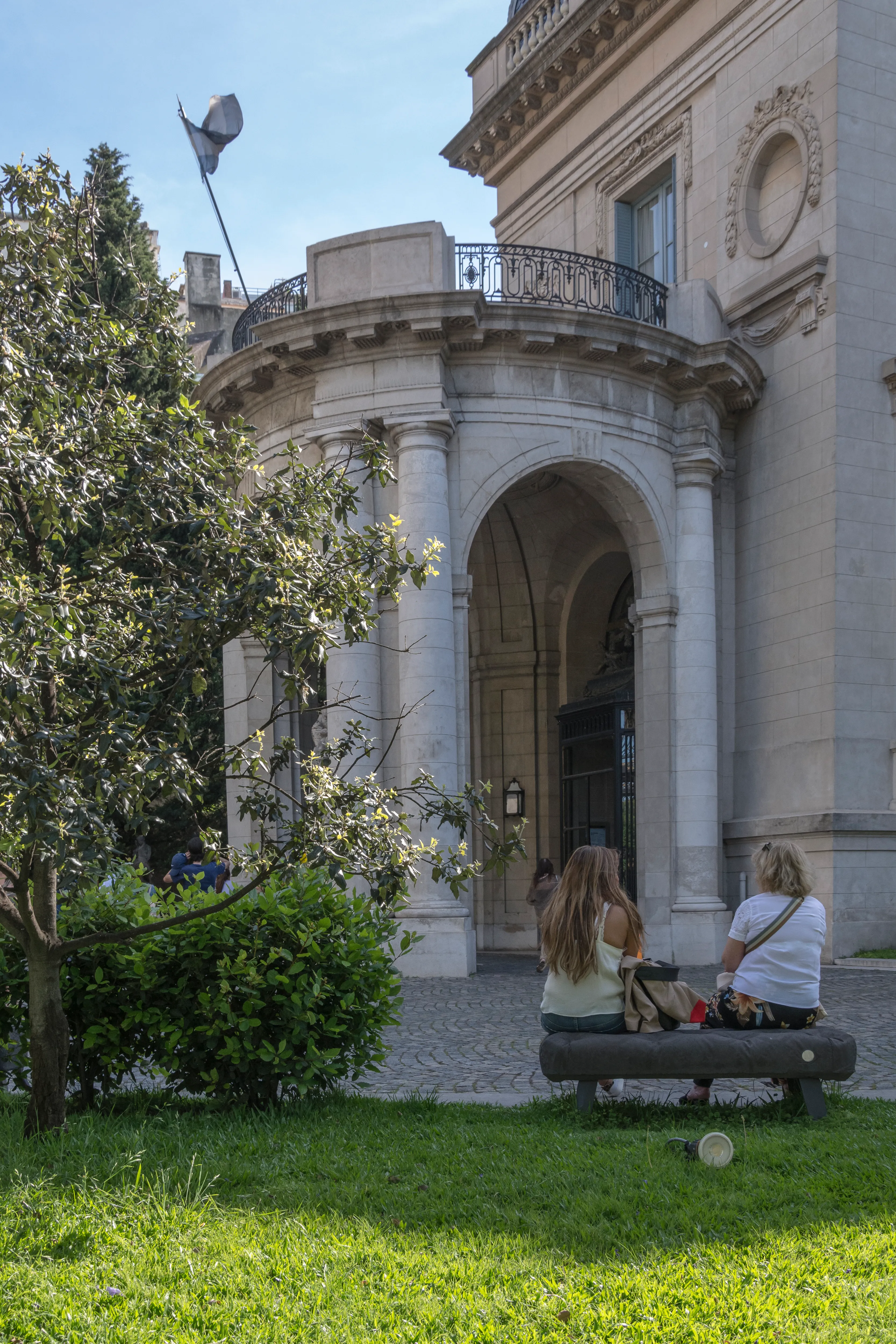 Dos personas sentadas en un banco frente a la fachada de piedra del Museo Nacional de Artes Decorativas, con un árbol y un césped verde en primer plano.