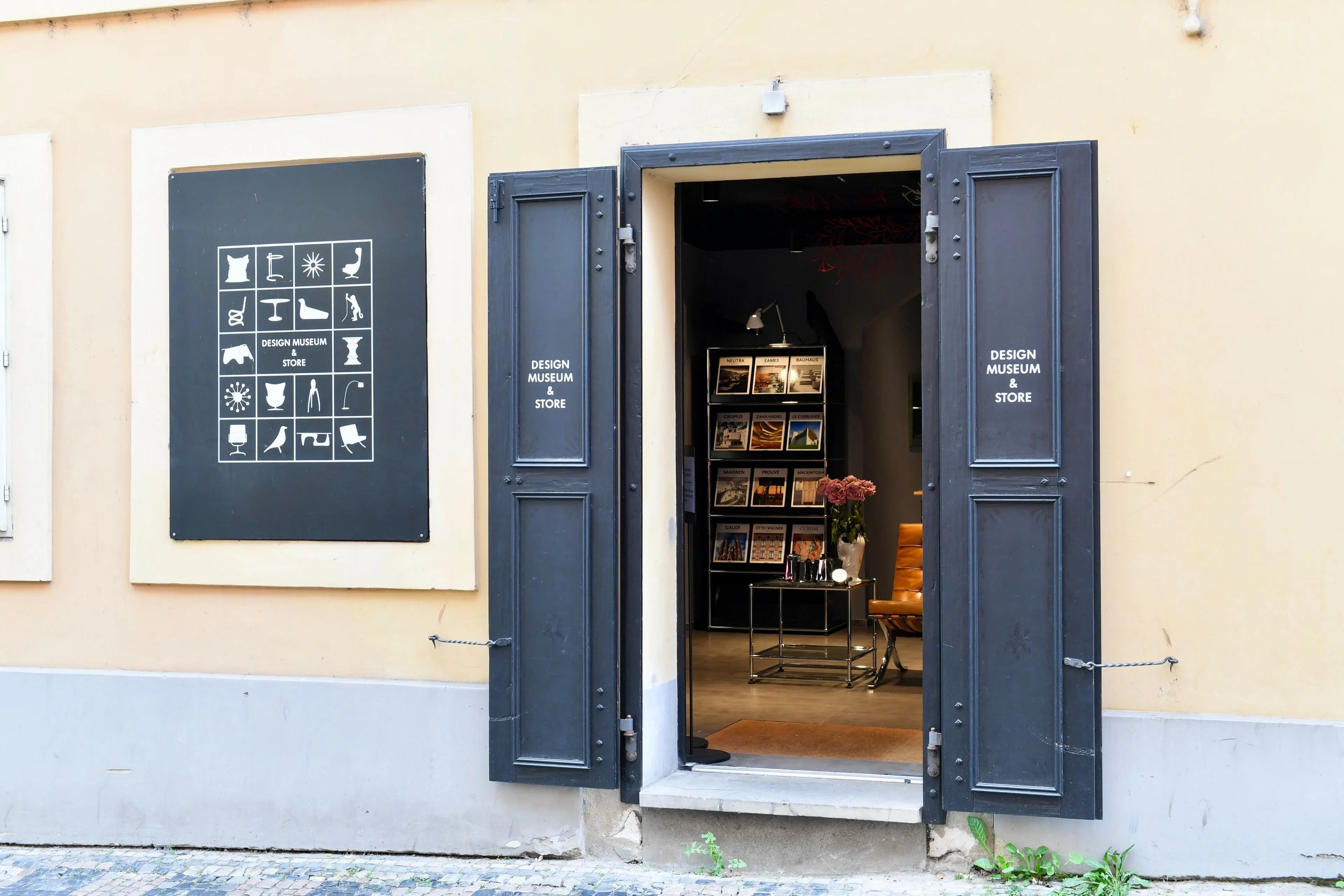 Open zwarte luiken aan een geel gebouw, die het interieur van het Design Museum Praag onthullen met boekenplanken en een vitrine.