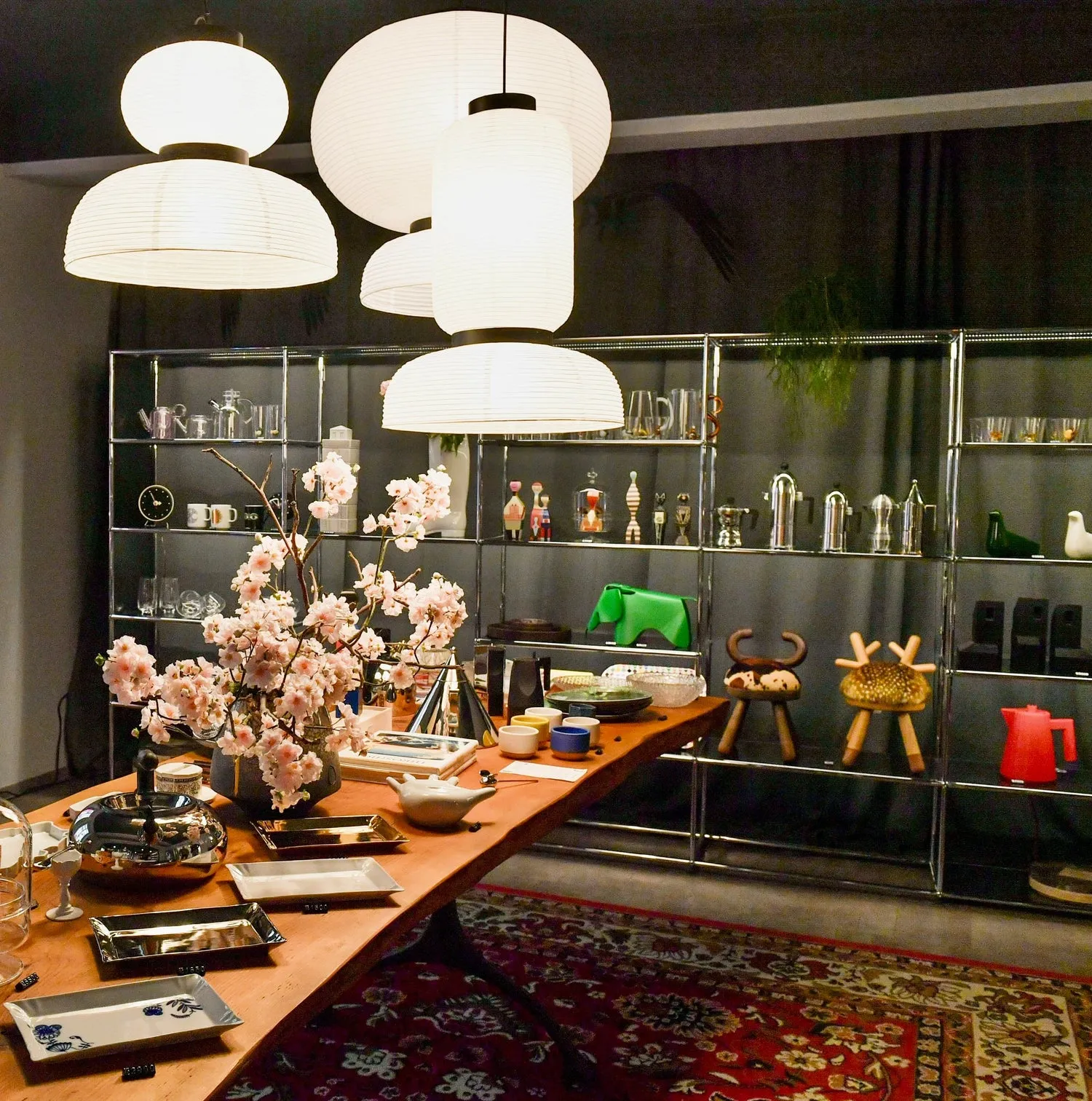 Tentoonstellingsruimte met een houten tafel waarop designobjecten, moderne hanglampen en glazen planken met diverse voorwerpen worden tentoongesteld