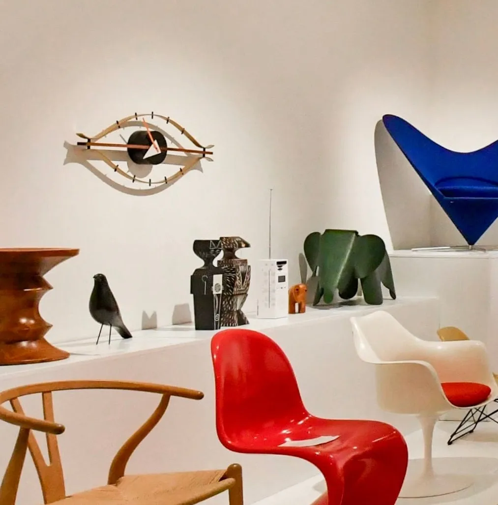 Moderne designobjecten tentoongesteld, waaronder een rode stoel, een blauwe stoel, een houten oogklok en een zwart vogelbeeld.