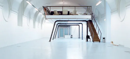 Un espacio interior moderno y blanco con un entresuelo soportado por vigas negras y una escalera de madera