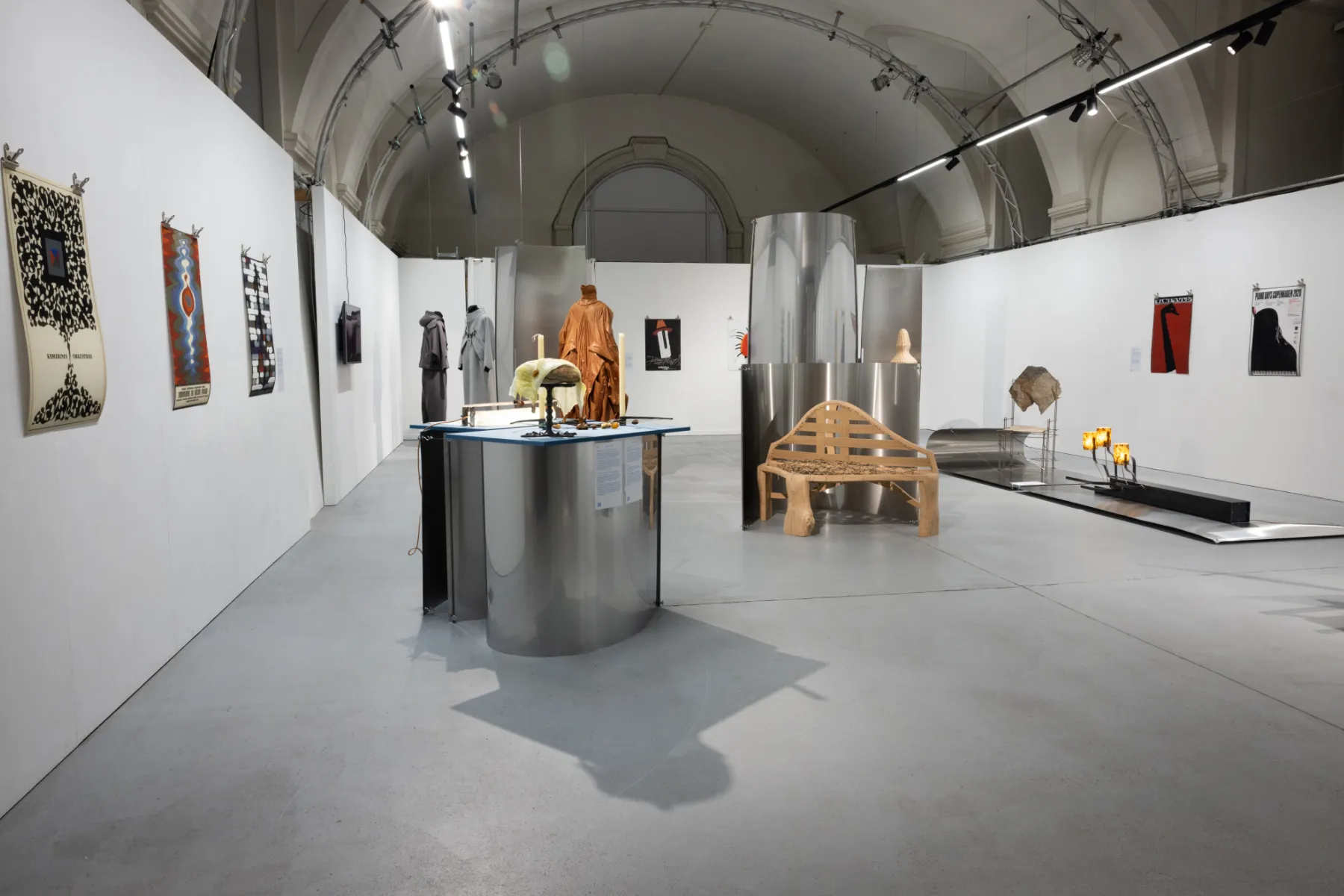 Interior de designforum Wien con un montaje de exposición que incluye una mesa metálica, un banco de madera y obras de arte en paredes blancas bajo un techo abovedado