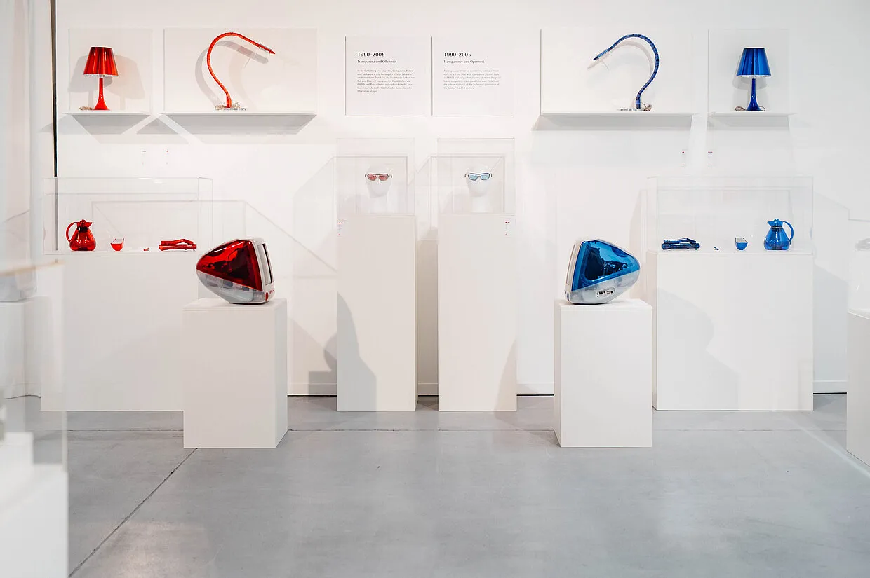 Objetos de diseño moderno que incluyen lámparas rojas y azules y piezas esculturales expuestas sobre pedestales blancos en un espacio de galería.