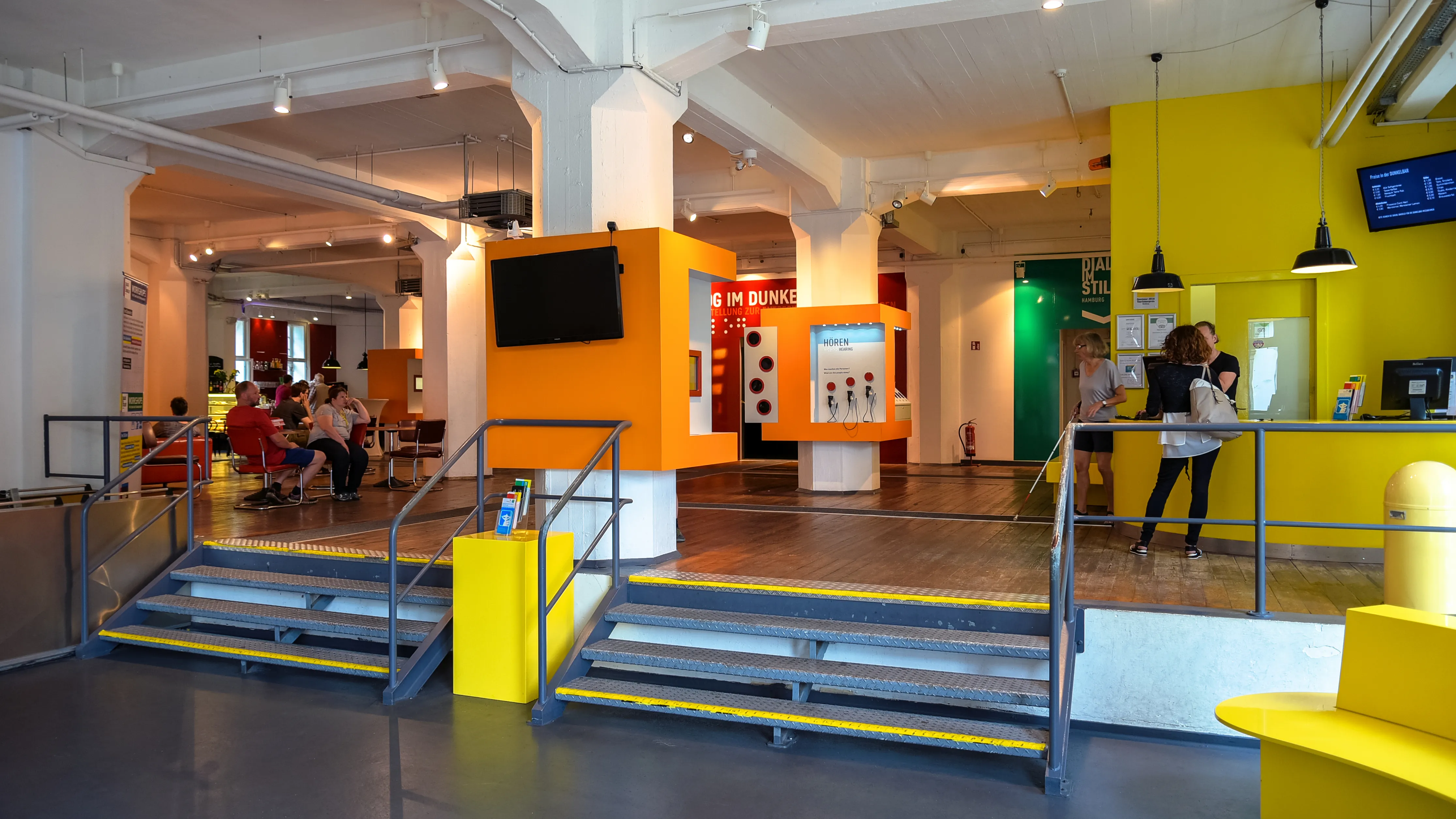 Interior de Dialog im Dunkeln con paredes amarillas y naranjas, una escalera y visitantes en un vestíbulo moderno