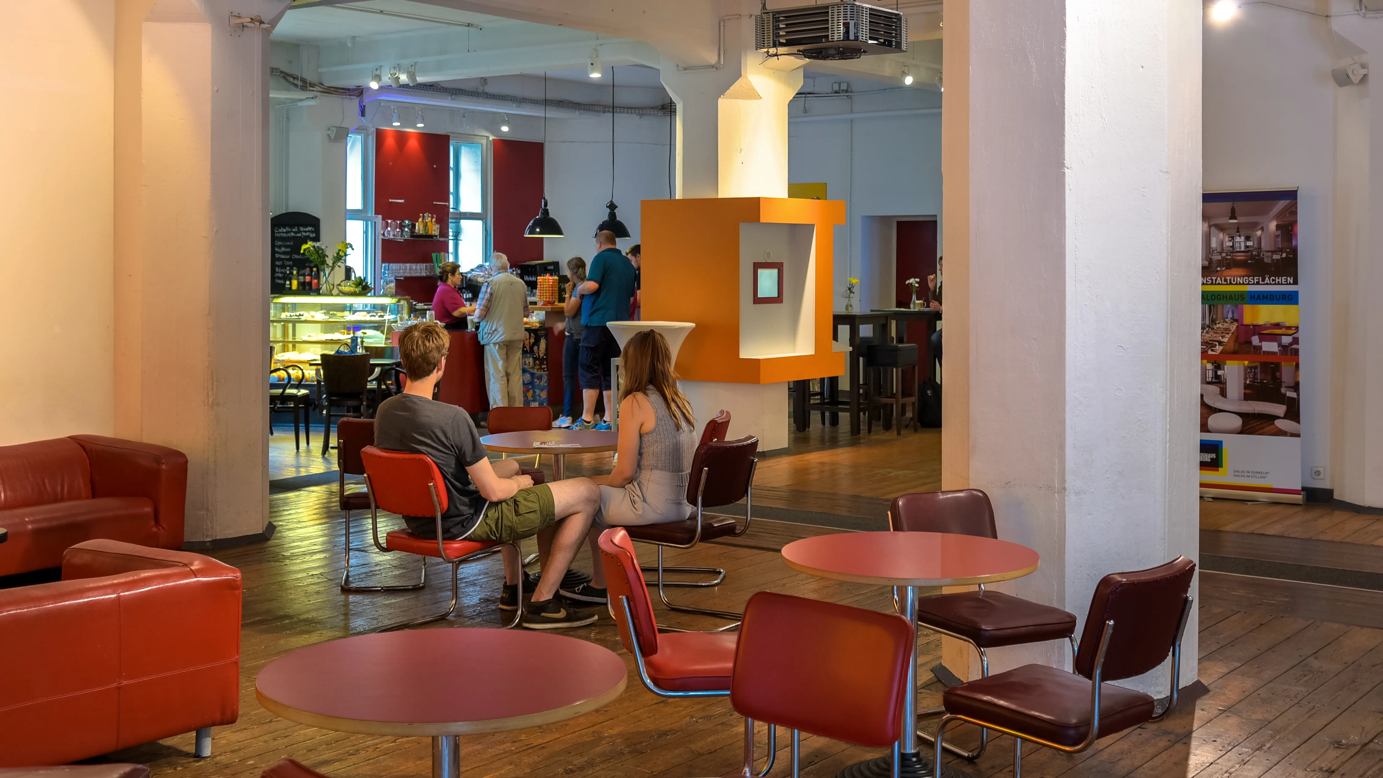 Sillas y mesas rojas en una zona lounge moderna con gente sentada y un fondo de luz brillante