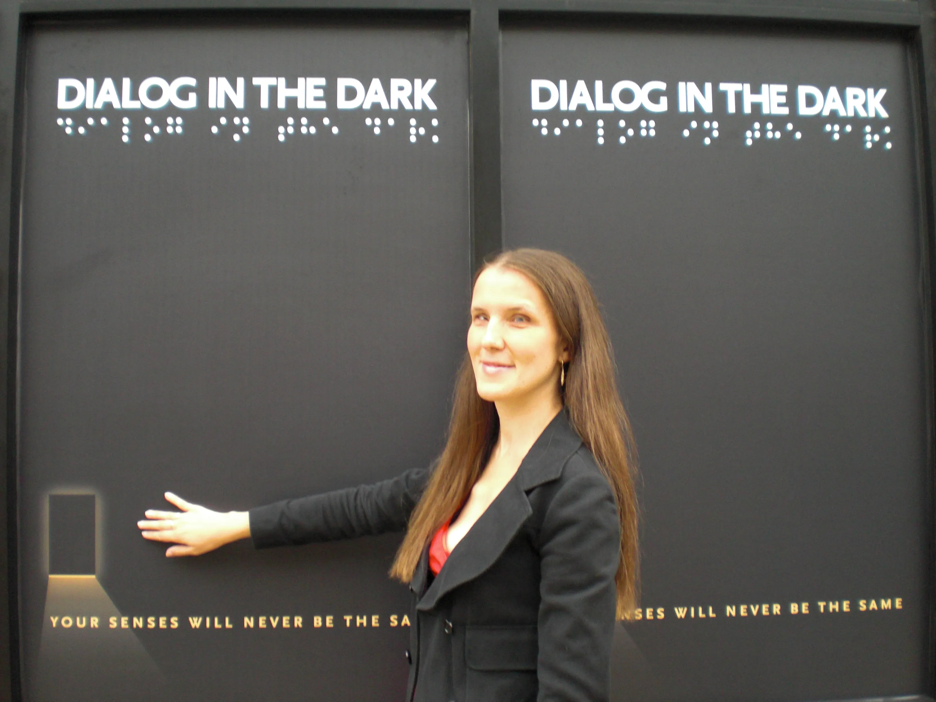 Una mujer con un blazer negro está junto a una pared negra con el texto 'DIALOG IN THE DARK' y braille, extendiendo su mano hacia un pequeño panel iluminado.