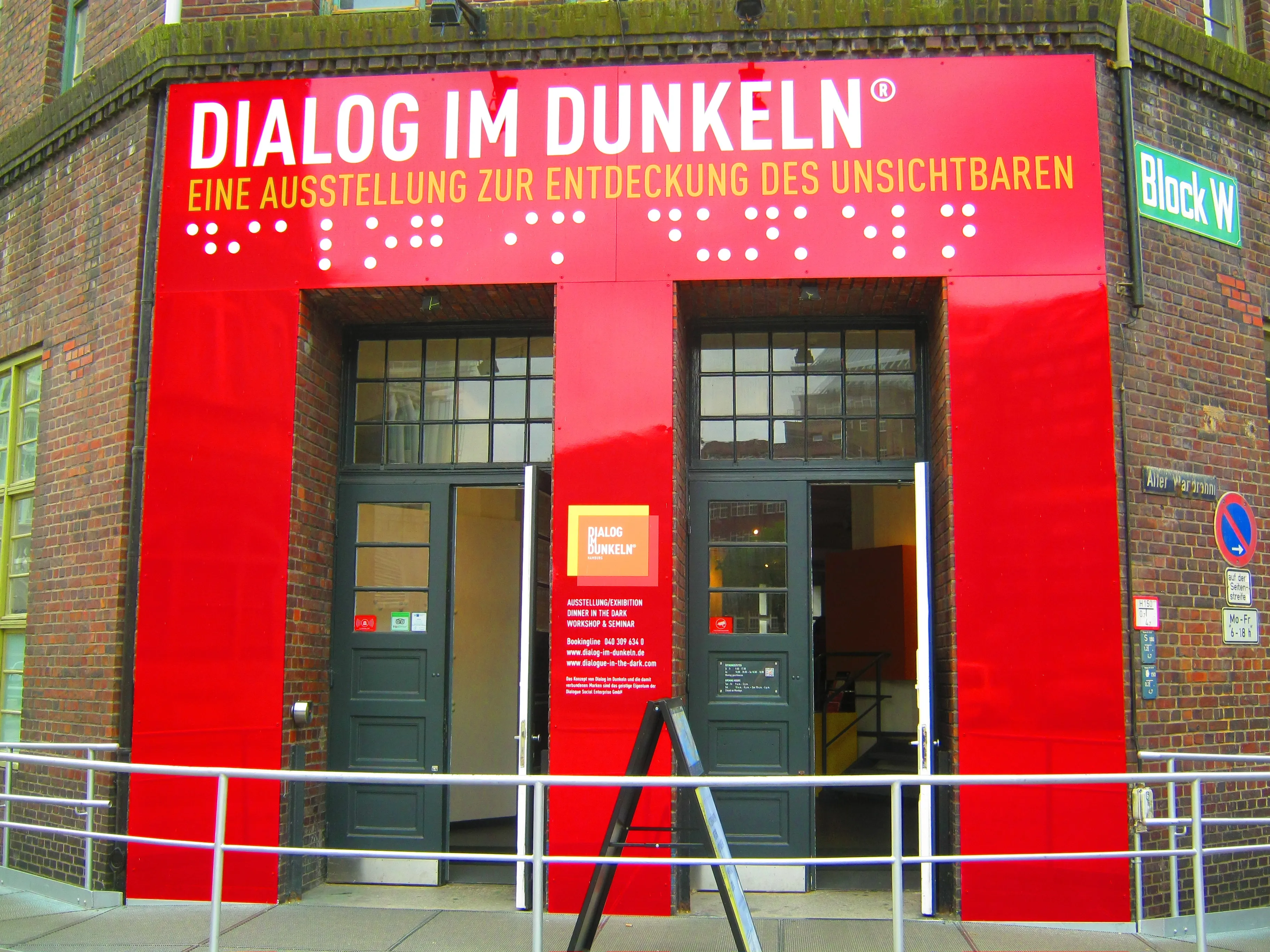 Fachada roja de Dialog im Dunkeln con texto blanco y braille, edificio de ladrillos, puertas de vidrio y una barandilla de metal delante