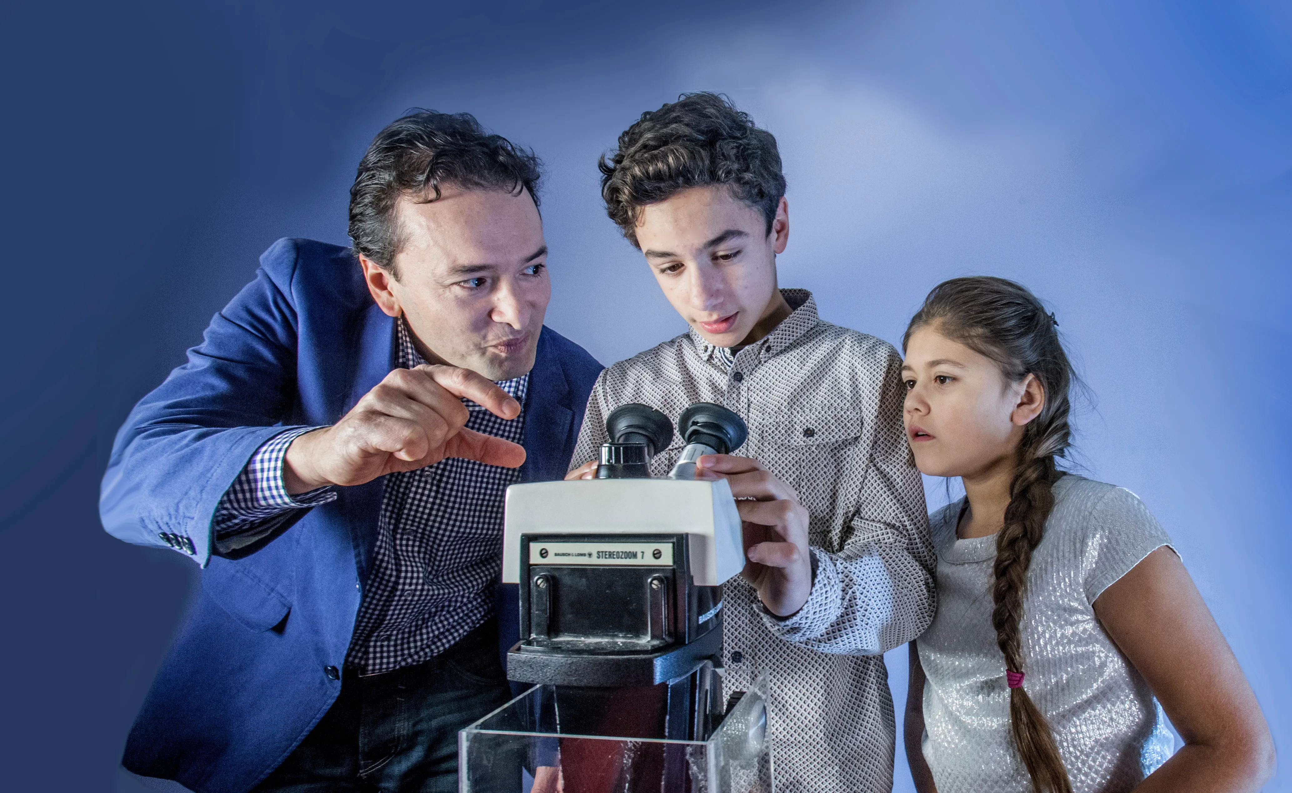 Un hombre y dos niños mirando a través de un microscopio binocular con un fondo azul.