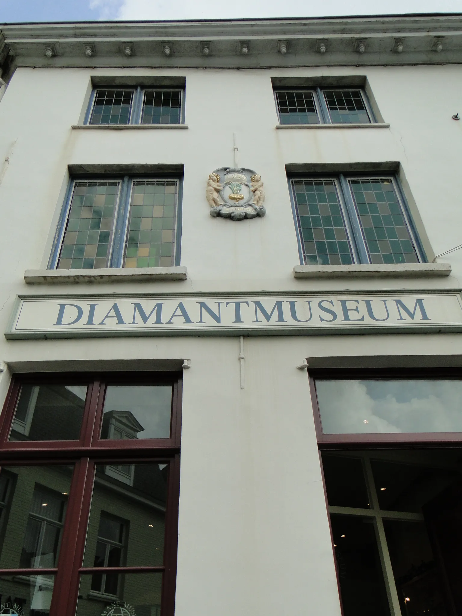 Fachada blanca de un edificio con un cartel grande que dice 'DIAMANTMUSEUM' y un escudo decorativo montado sobre él, con varias ventanas de marco oscuro distribuidas en dos plantas.