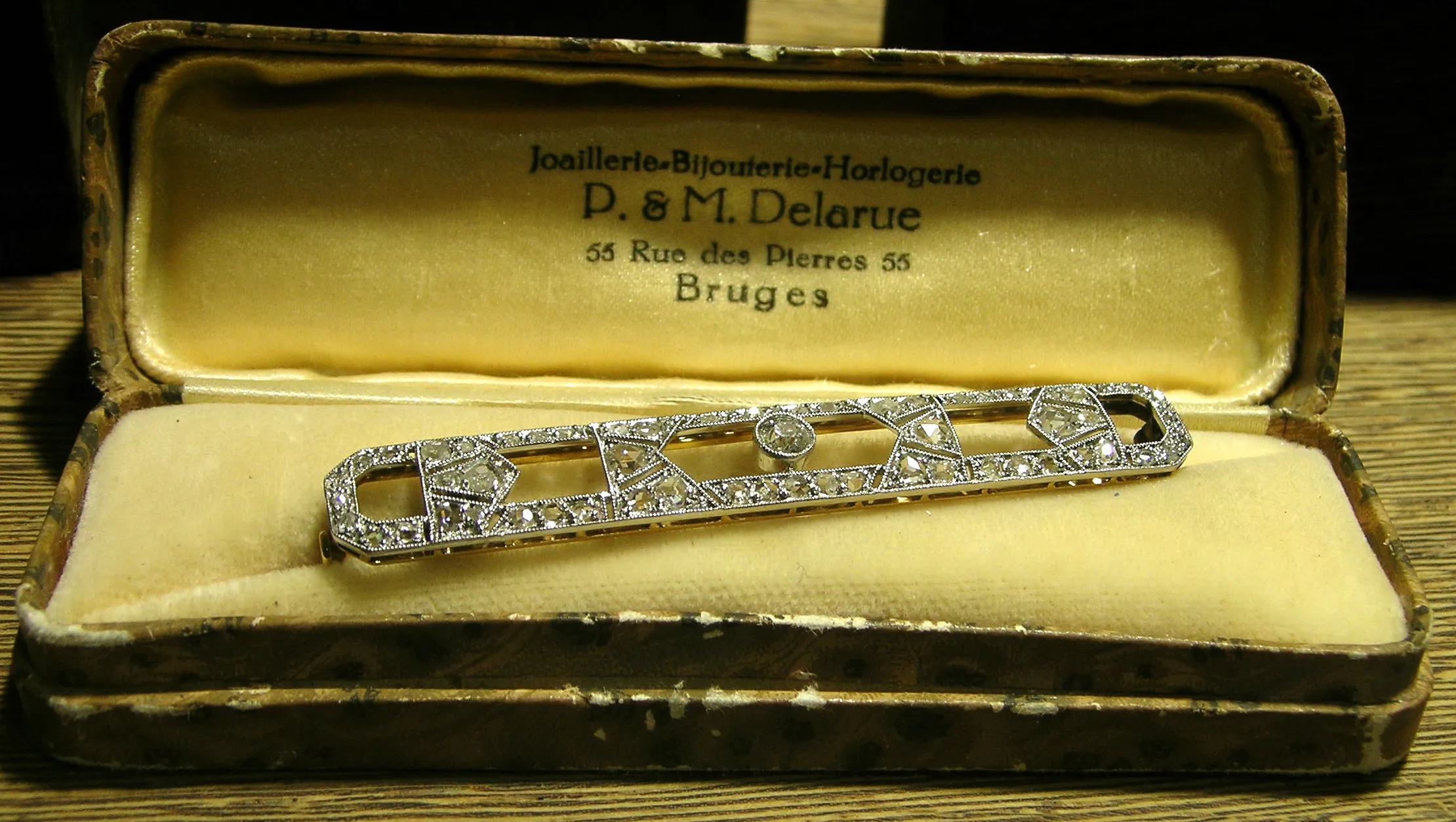 Una pulsera de diamantes descansando dentro de un joyero vintage abierto, con una tapa forrada de terciopelo grabada con "P. & M. Delarue, 55 Rue des Pierres, Bruges".
