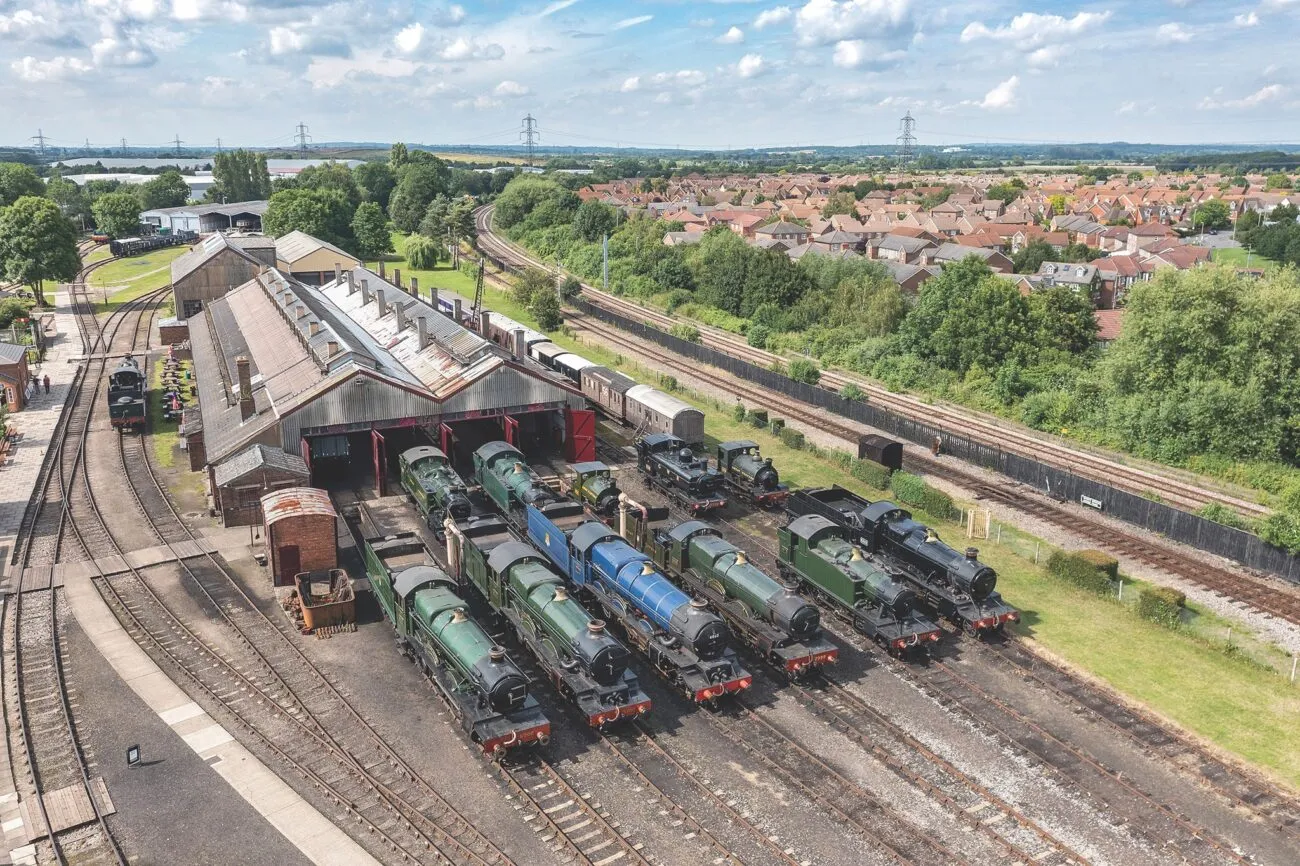 Widok z lotu ptaka na kilka parowozów stojących na torach przed dużą parowozownią w Didcot Railway Centre, z dzielnicą mieszkalną w tle.