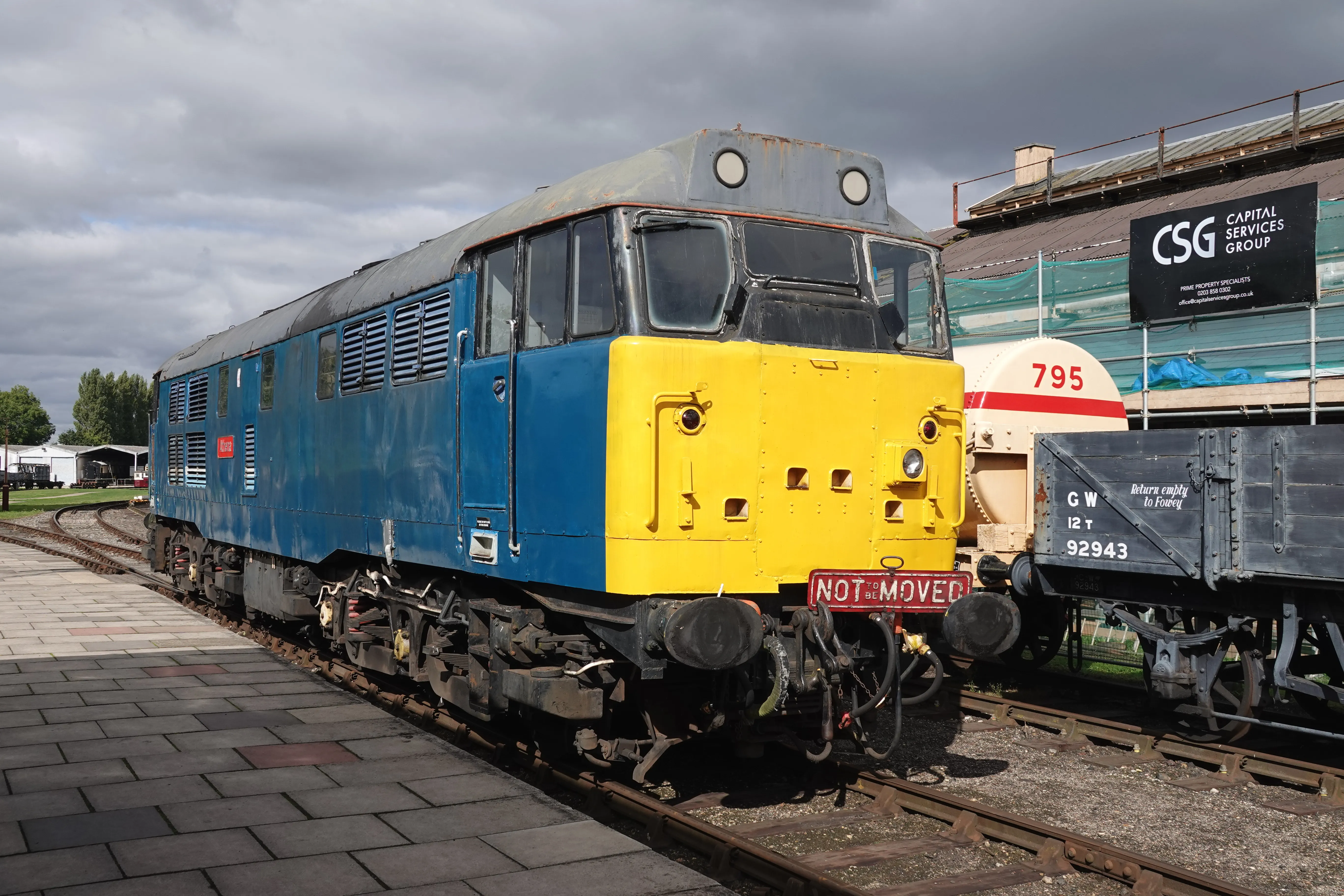 Niebiesko-żółta lokomotywa spalinowa z czerwoną tabliczką „NOT MOVED” na przedniej belce zderzakowej, zaparkowana na torach obok wagonu towarowego w Didcot Railway Centre.