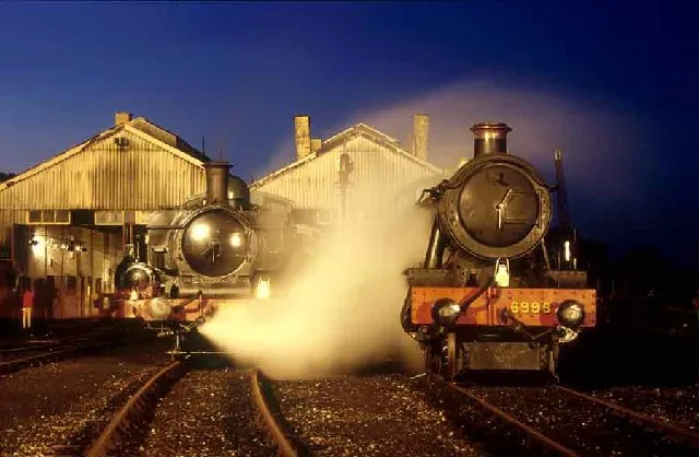 Dwie lokomotywy parowe o zmierzchu z jasną parą unoszącą się między nimi przed pofalowanymi halami parowozowni w Didcot Railway Centre, na tle ciemnoniebieskiego nocnego nieba.