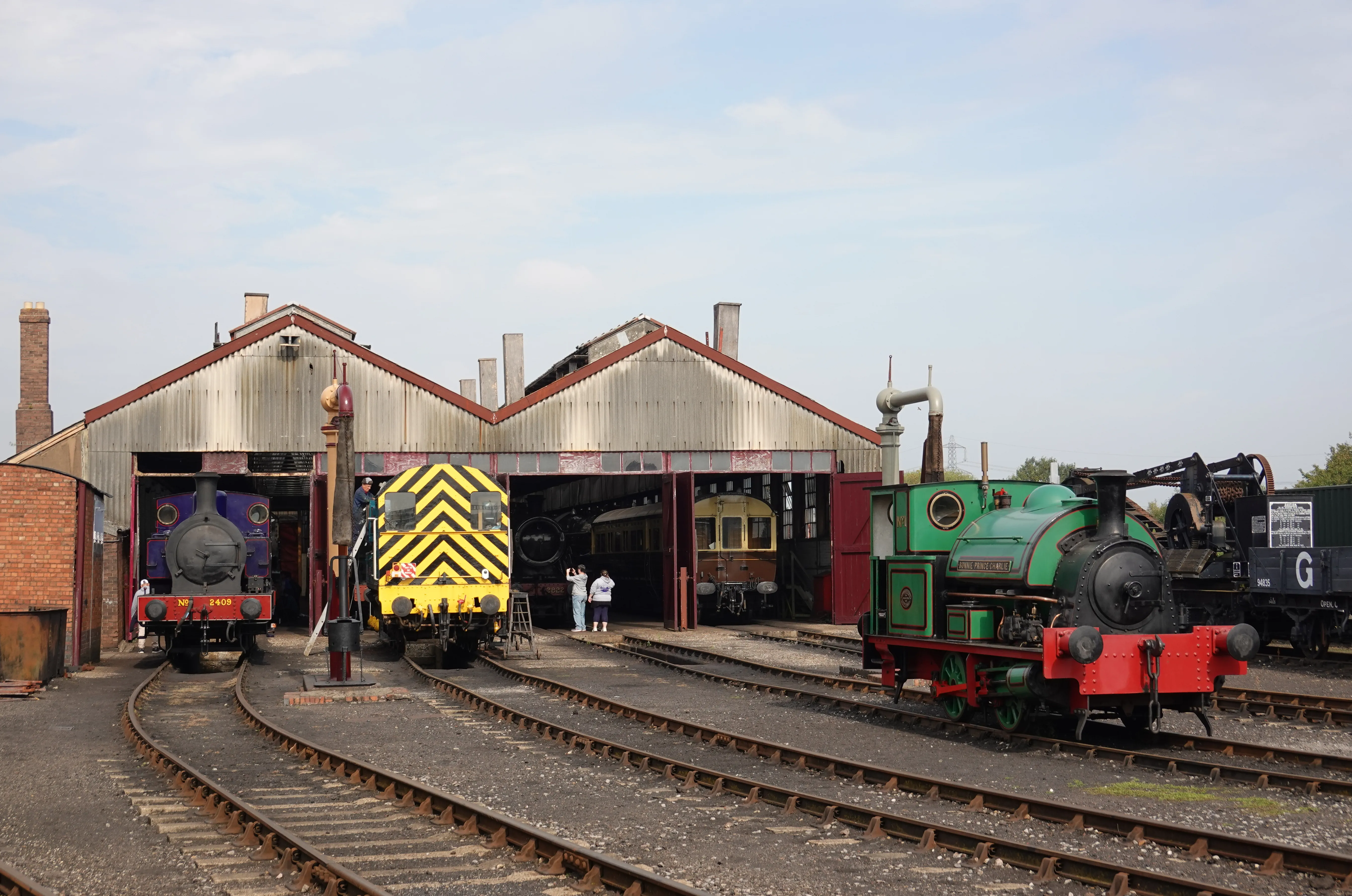 Zabytkowe lokomotywy, w tym zielona maszyna parowa i żółta lokomotywa w chevronowy wzór, zaparkowane przed parowozownią w Didcot Railway Centre