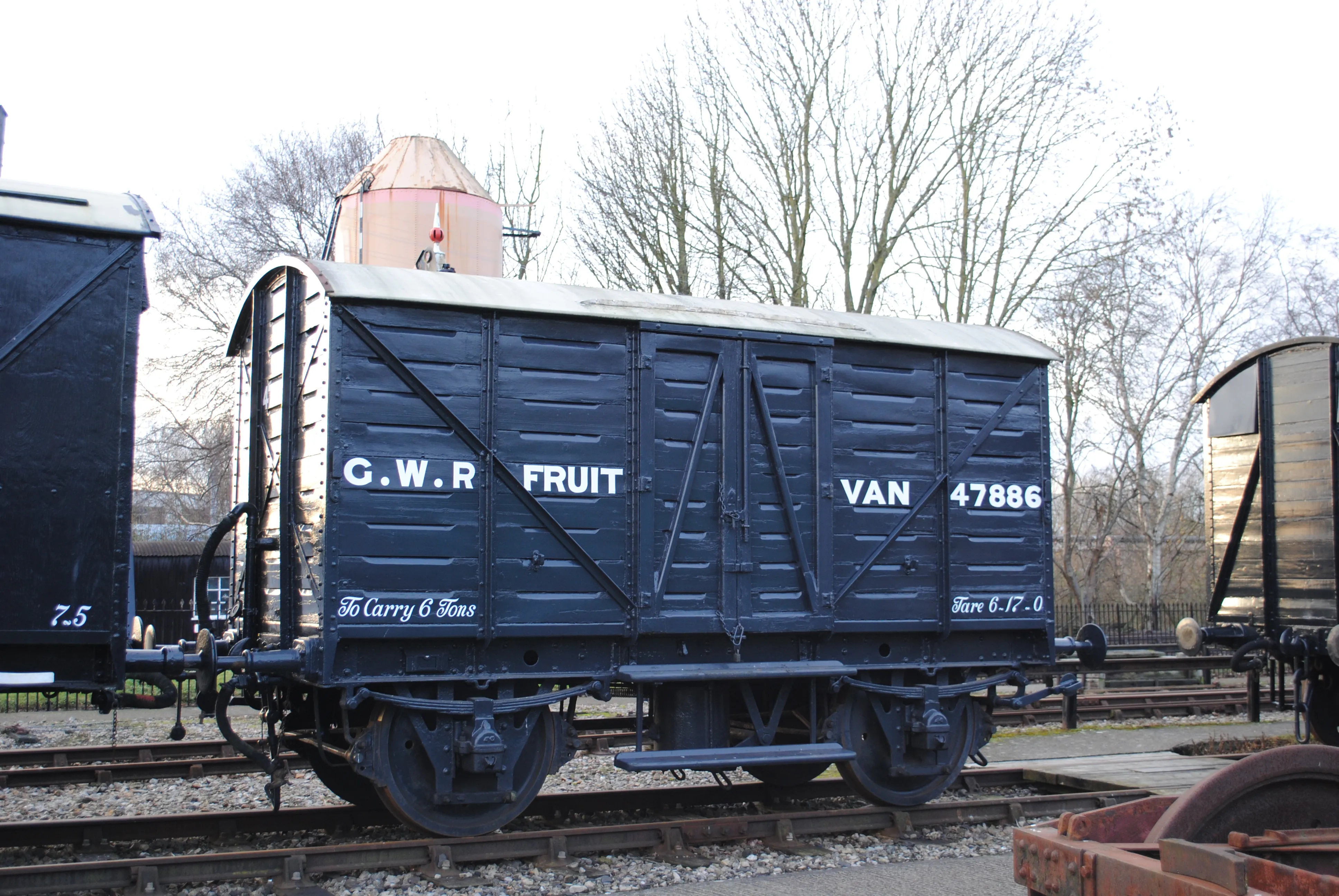 Ciemnoniebieski wagon towarowy G.W.R. Fruit Van nr 47886 stojący na torach na zewnątrz, z nagimi drzewami w tle.