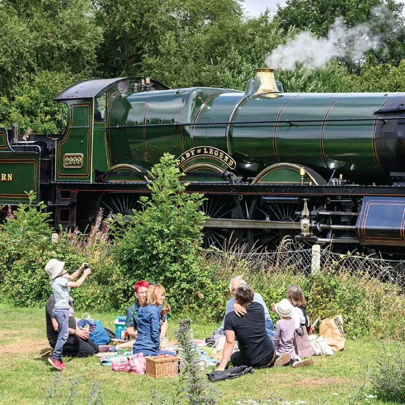 Rodziny piknikujące na trawie przed zieloną lokomotywą parową GWR o numerze 2000 z mosiężną tabliczką znamionową, z unoszącą się parą i drzewami w tle