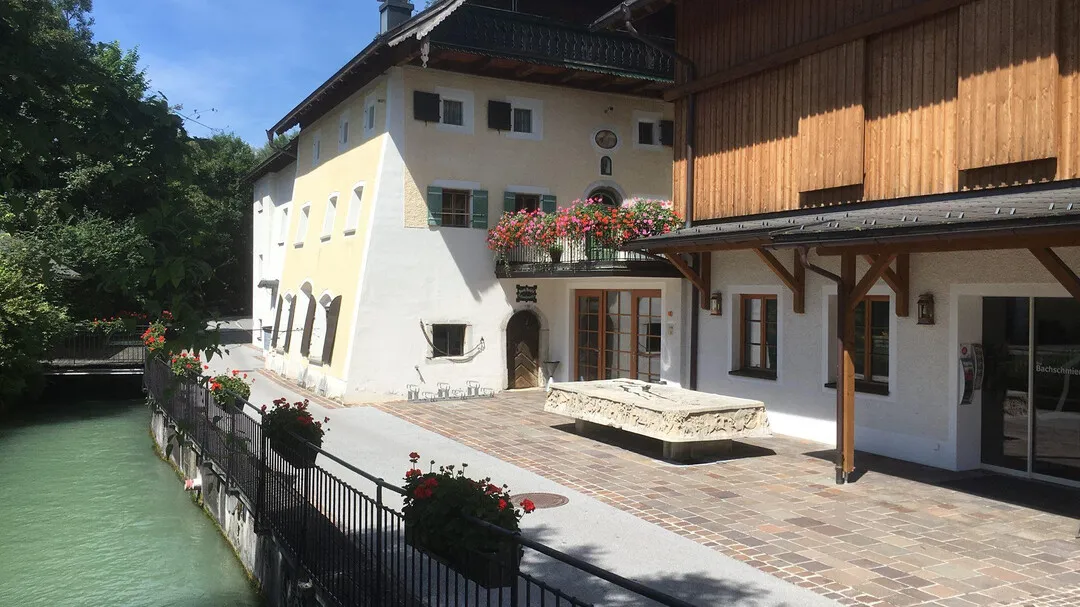 Edificio blanco y de madera con jardineras en los balcones junto a un canal verde, que cuenta con un patio pavimentado con una pila de piedra.